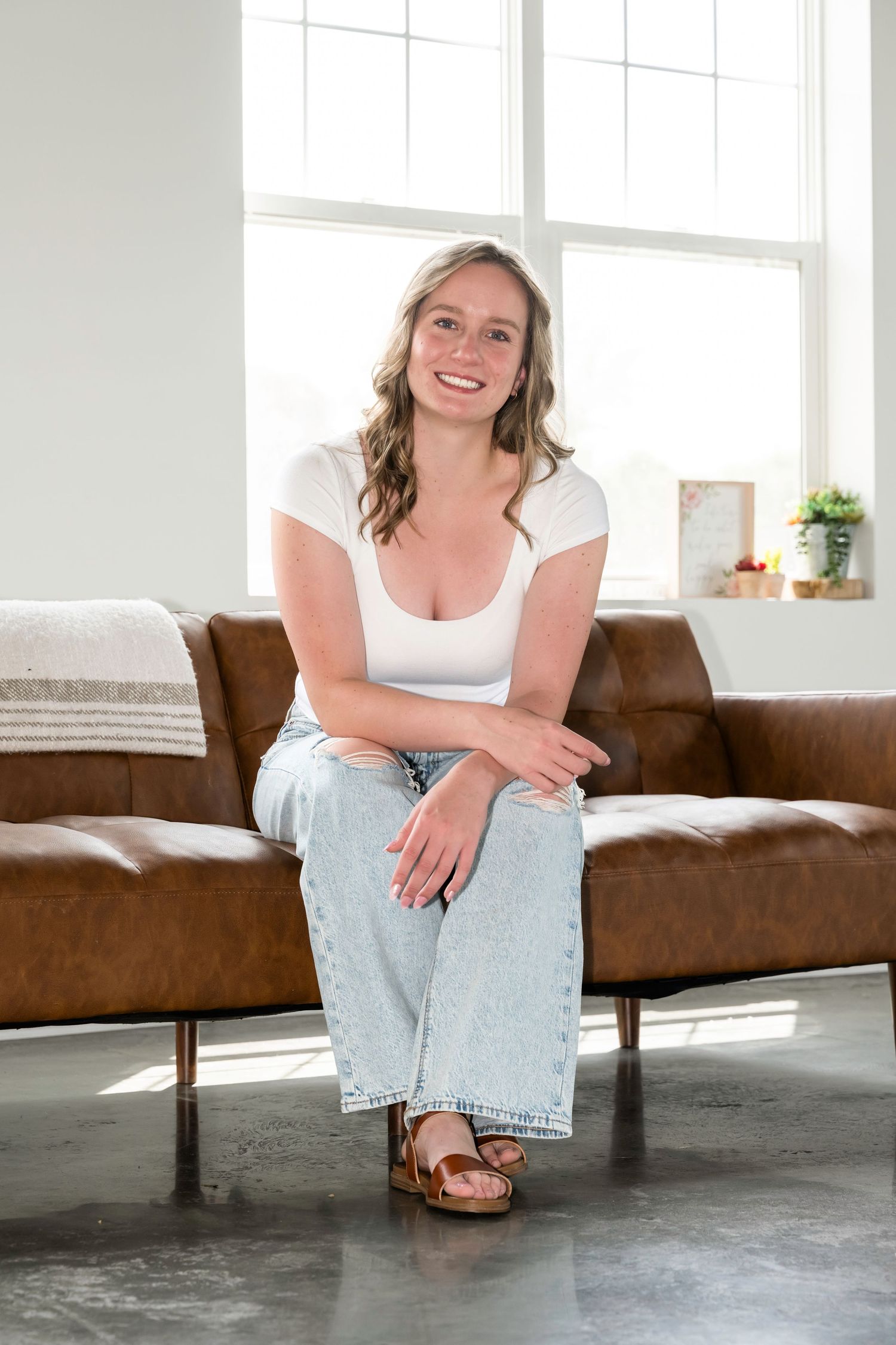 Smiling young woman in white top and jeans sitting on brown leather couch by bright window.