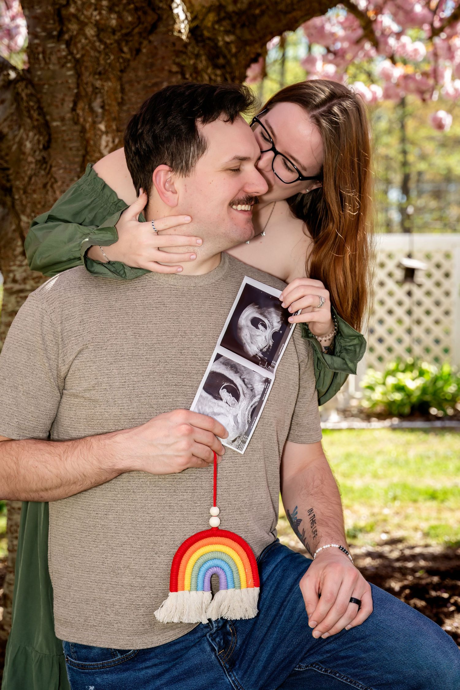 Happy couple sharing a tender moment outdoors, holding ultrasound photos and a rainbow decoration, announcing their pregnancy.