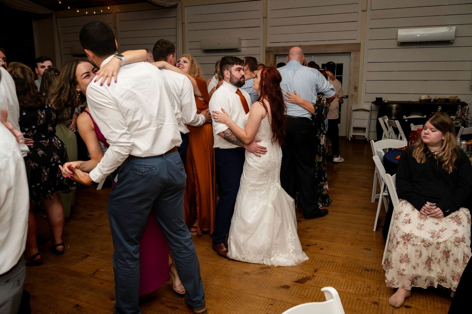 Bride and groom dance together with wedding guests on the reception dance floor in a rustic venue.