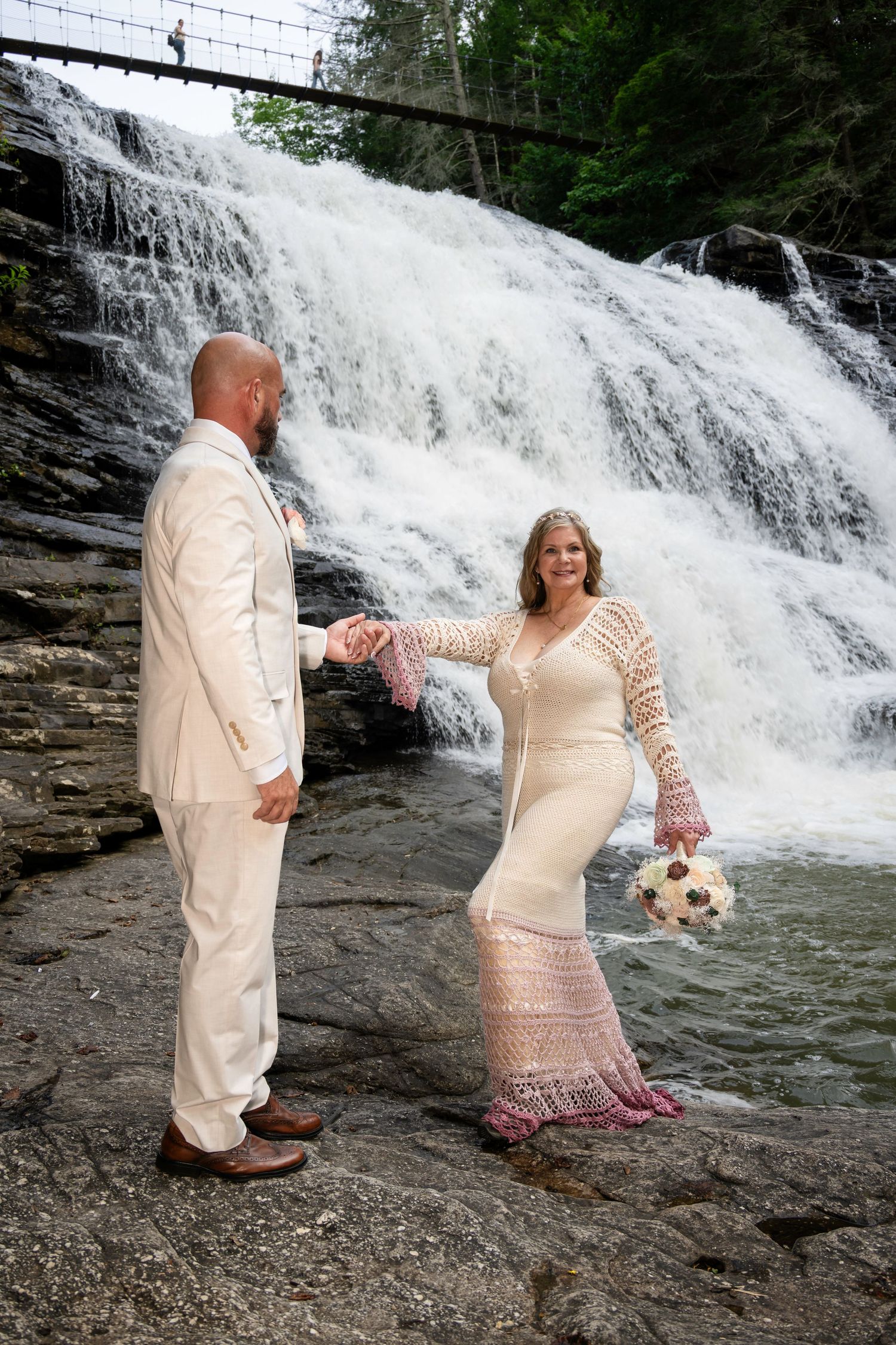 Bride and groom hold hands before a stunning waterfall backdrop, she in a lace gown, he in a white suit.
