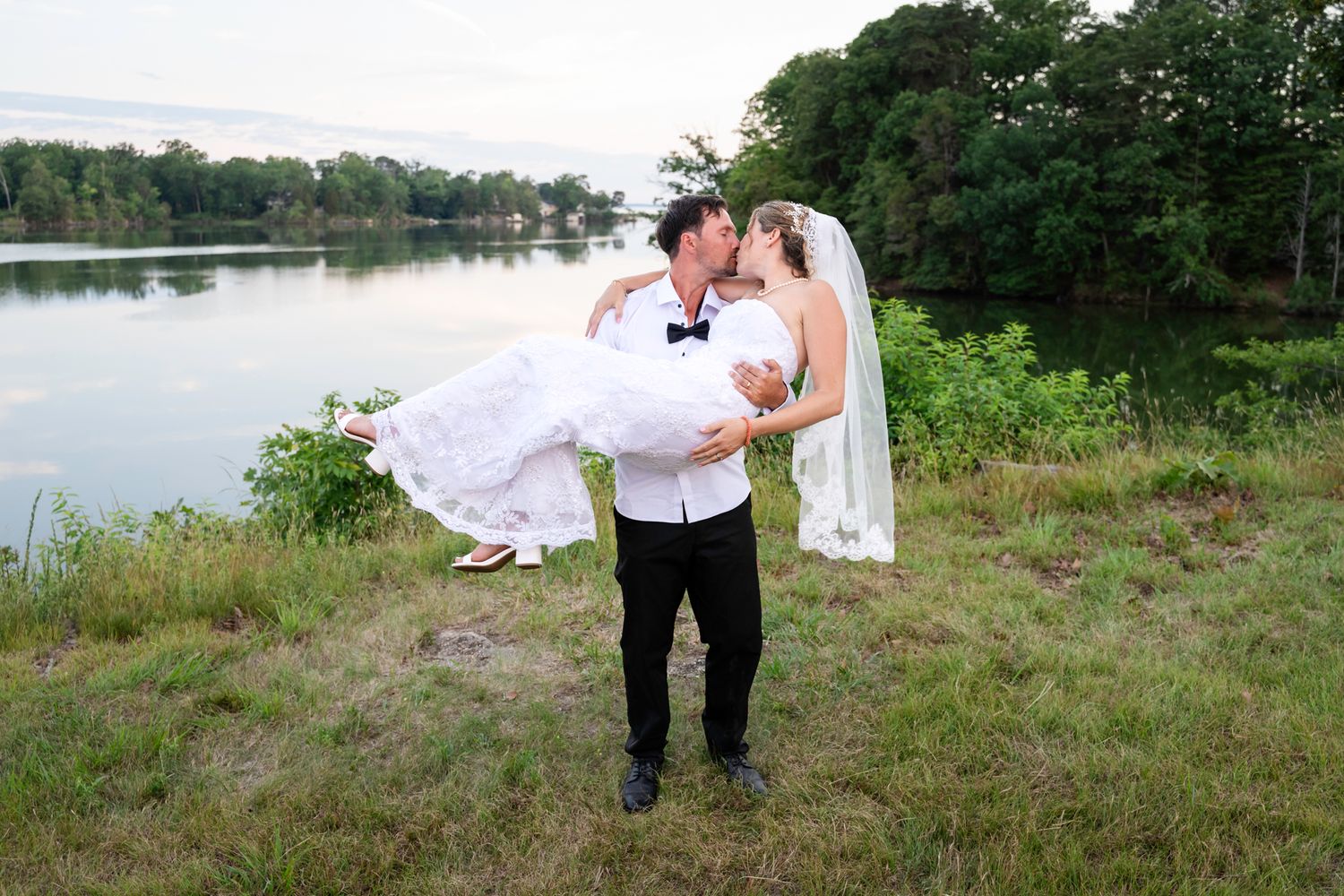 Groom carries bride in white wedding dress near a serene lake surrounded by lush green trees at outdoor wedding.