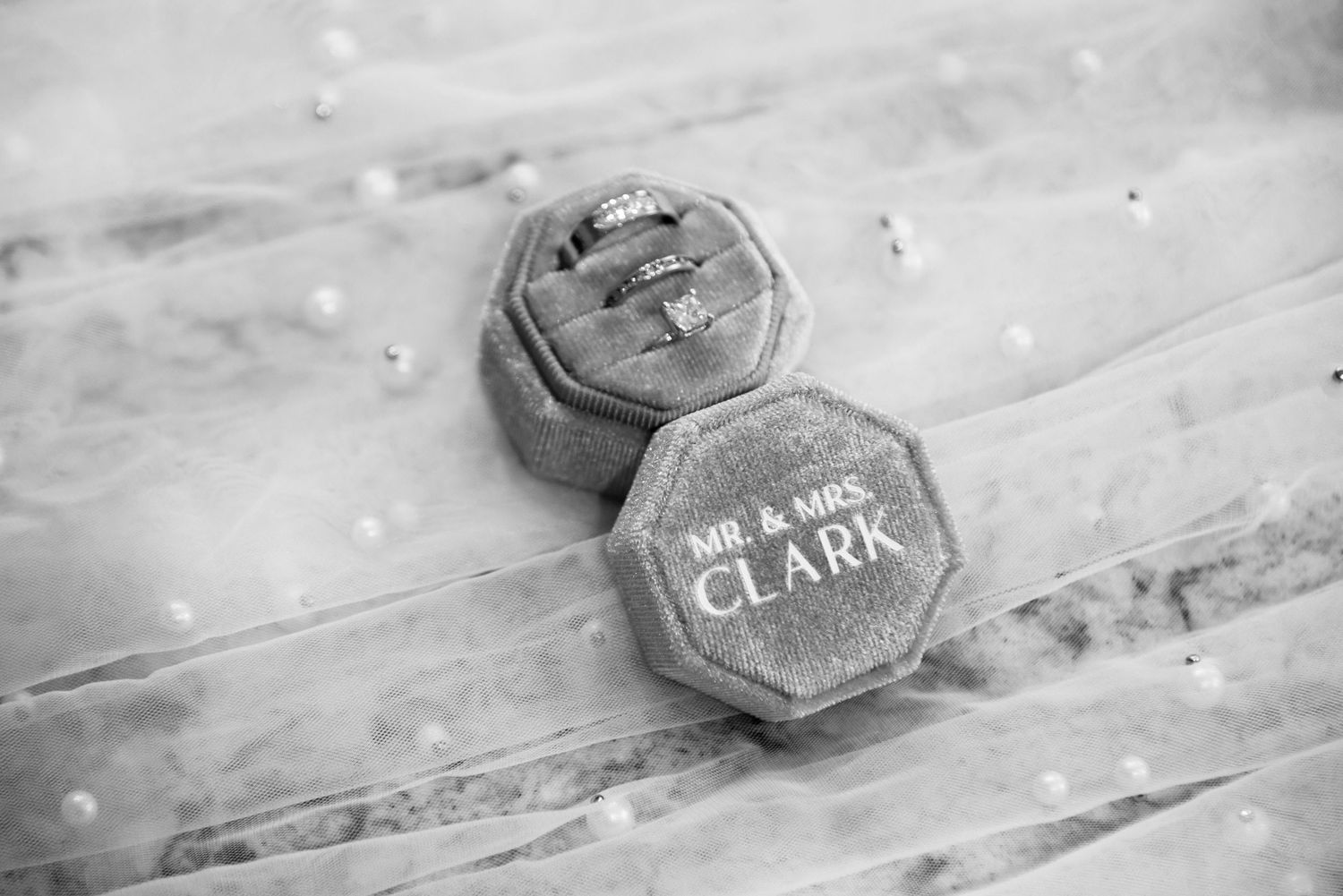 Black and white photo of Mr. & Mrs. Clark wedding ring box with elegant diamond rings on lace fabric.