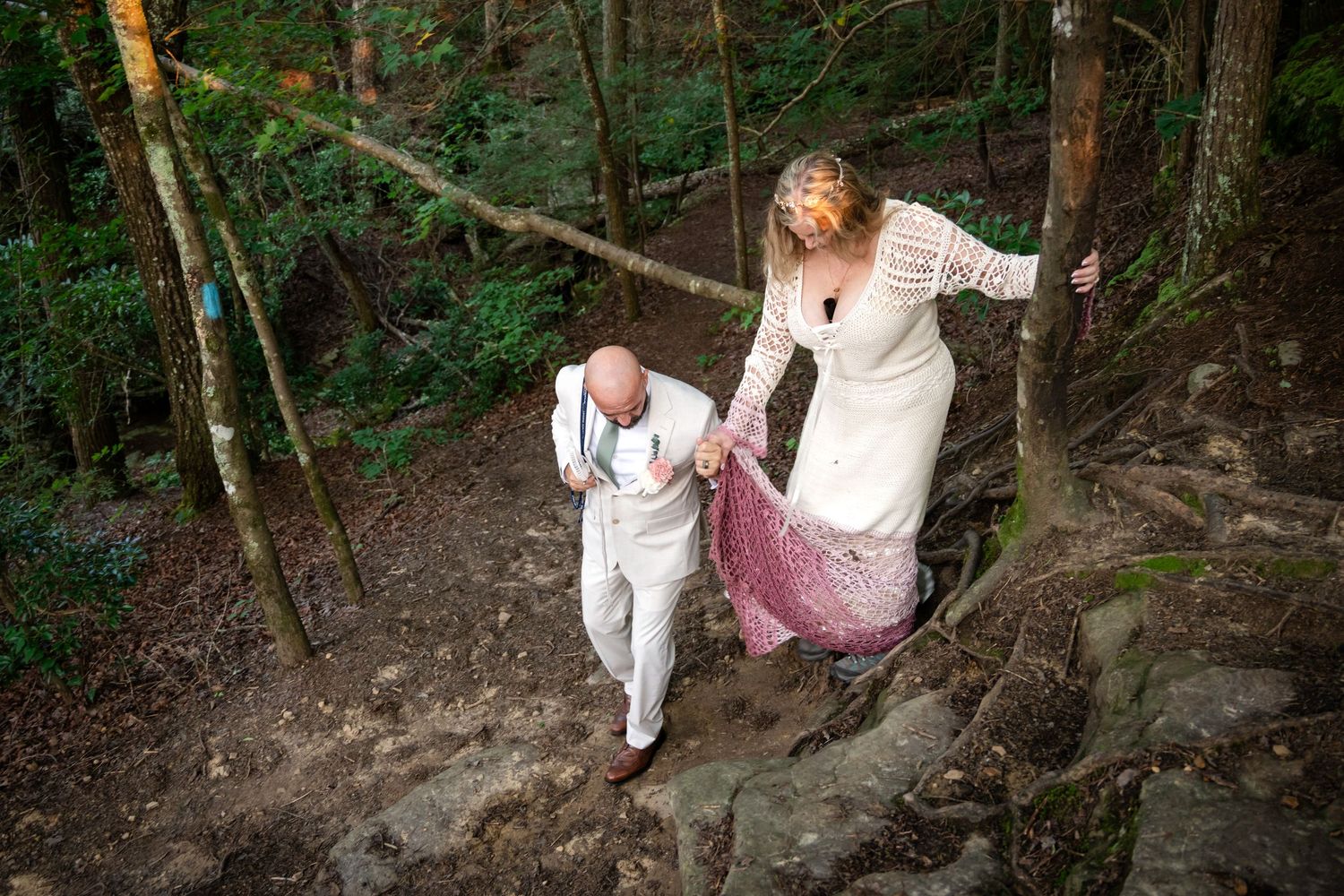 Bride and groom walking up rocky forest path, woman in white lace dress holding train, man in white suit leading the way.
