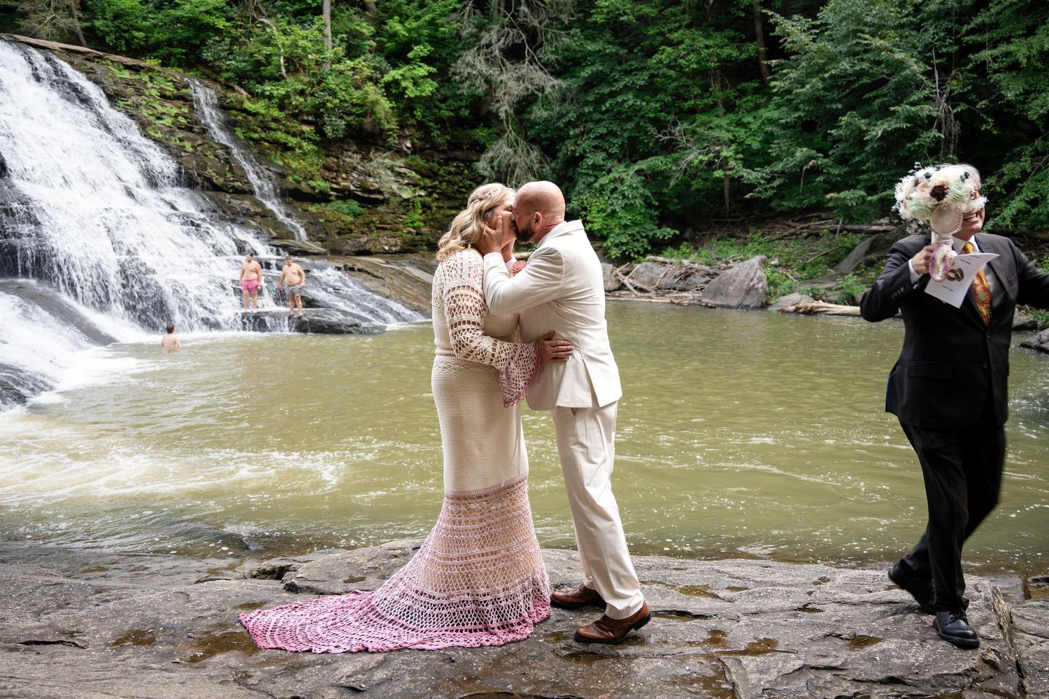 Couple sharing a kiss during outdoor wedding ceremony beside a scenic waterfall, officiant holding book nearby.