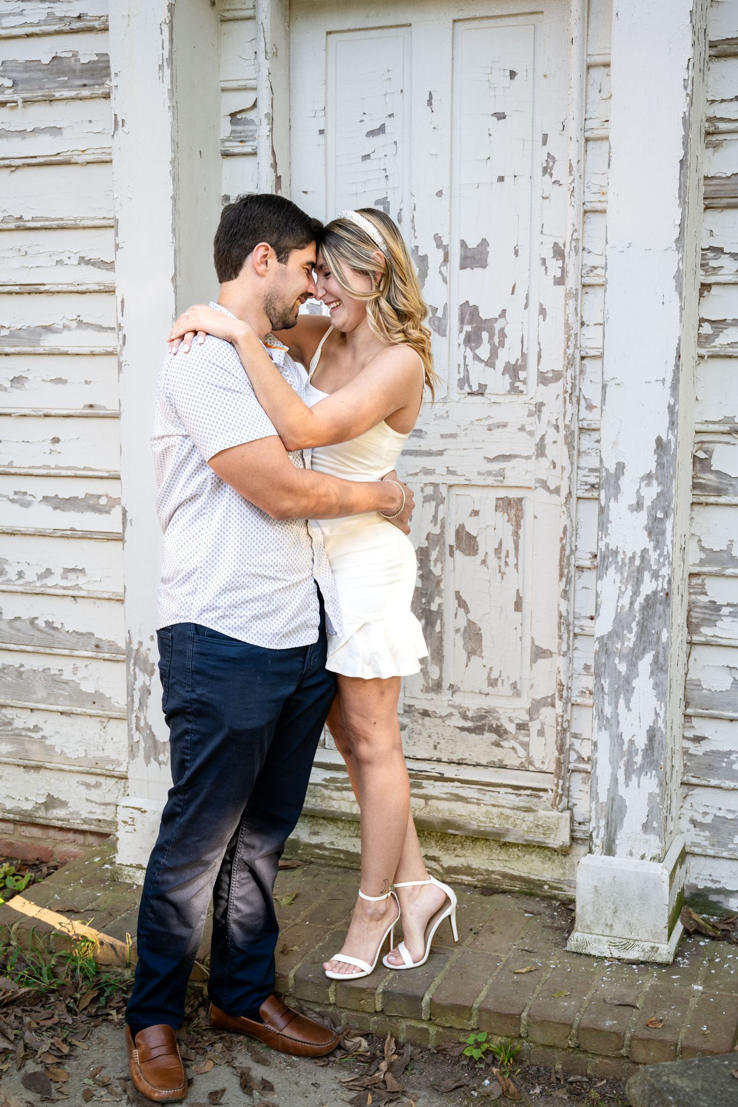 Couple embracing and touching foreheads romantically in front of a rustic white peeling paint door outdoors.