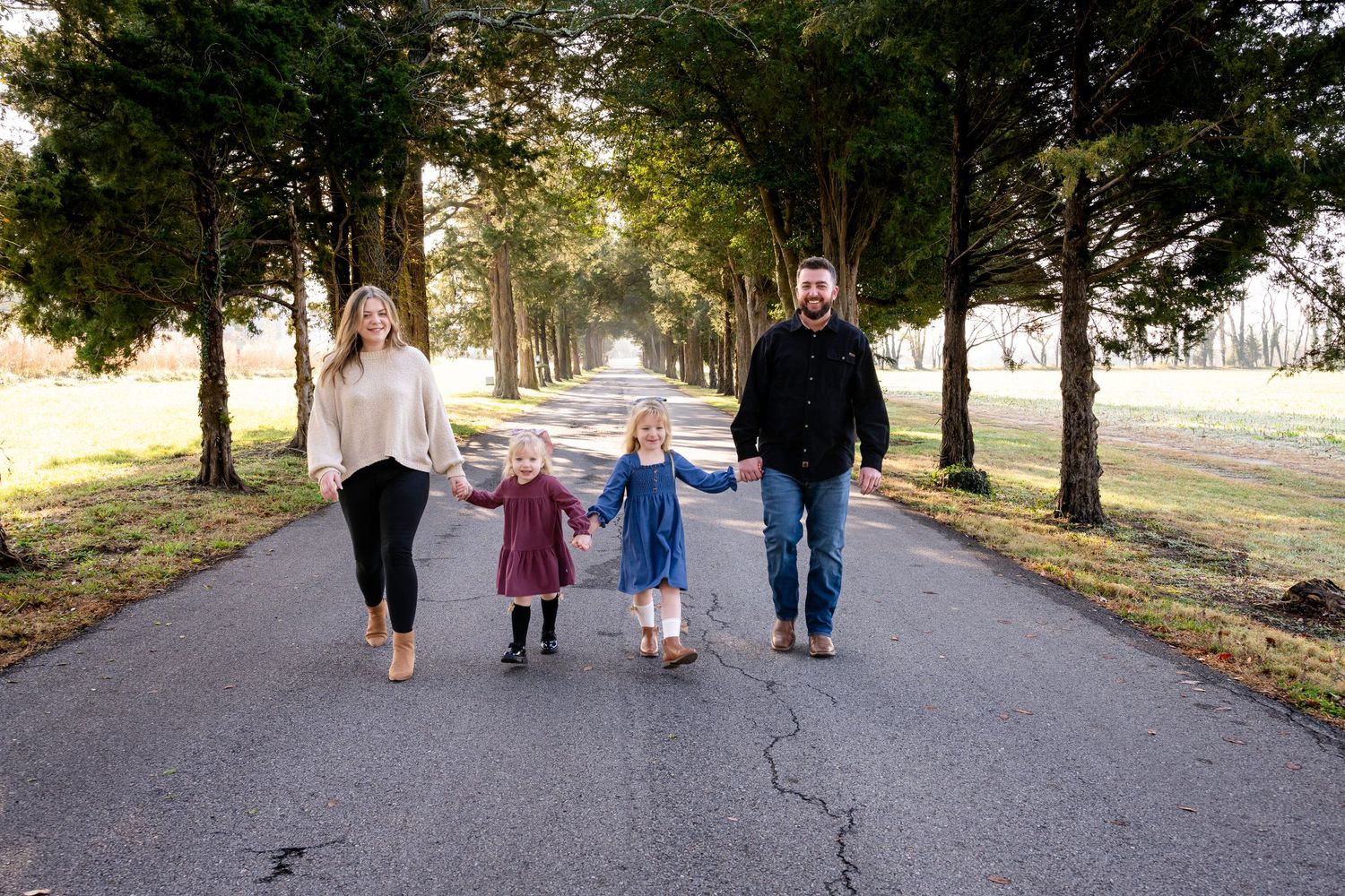 Family of four holding hands walking down a tree-lined road during a golden hour outdoor photo session.