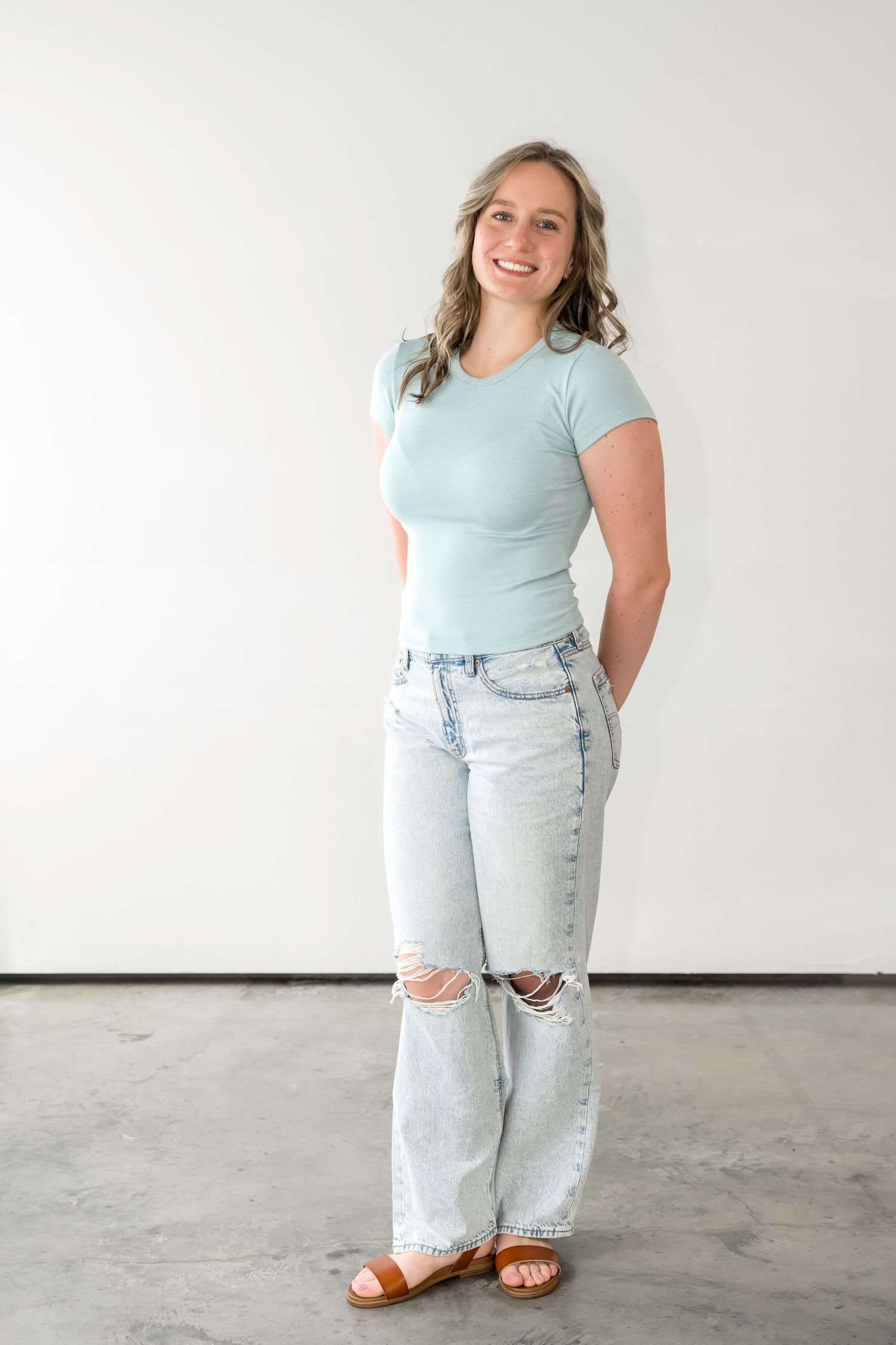 Woman smiling in mint green tee, ripped light wash wide-leg jeans, and tan sandals against white wall.
