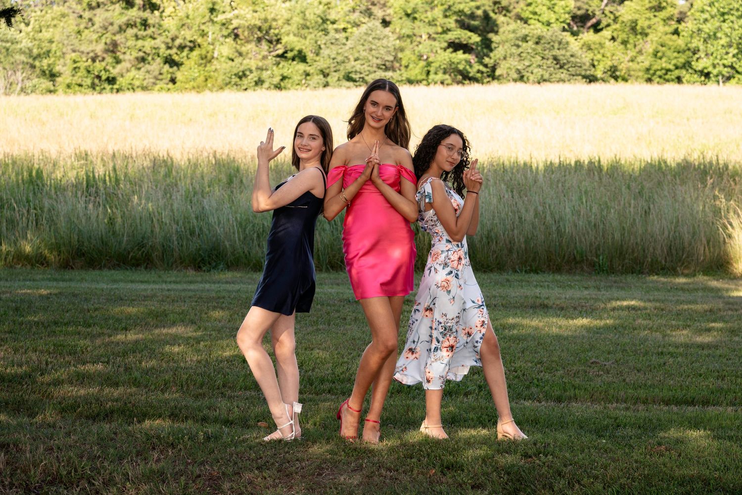 Three women in colorful summer dresses pose playfully on a green lawn with tall grass in the background.