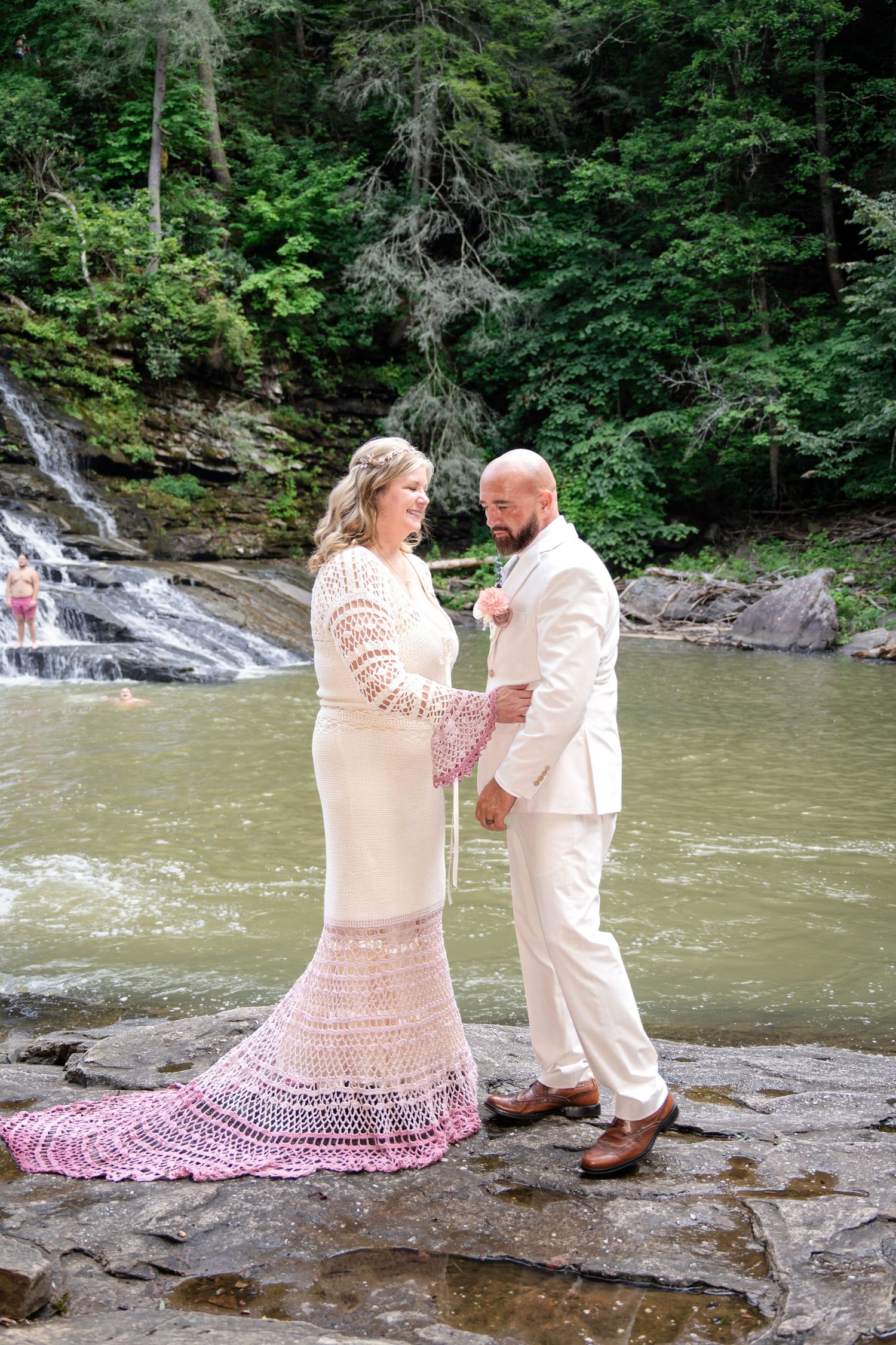 Bride in white crochet gown and groom in cream suit share a moment on rocks beside a waterfall in lush forest.