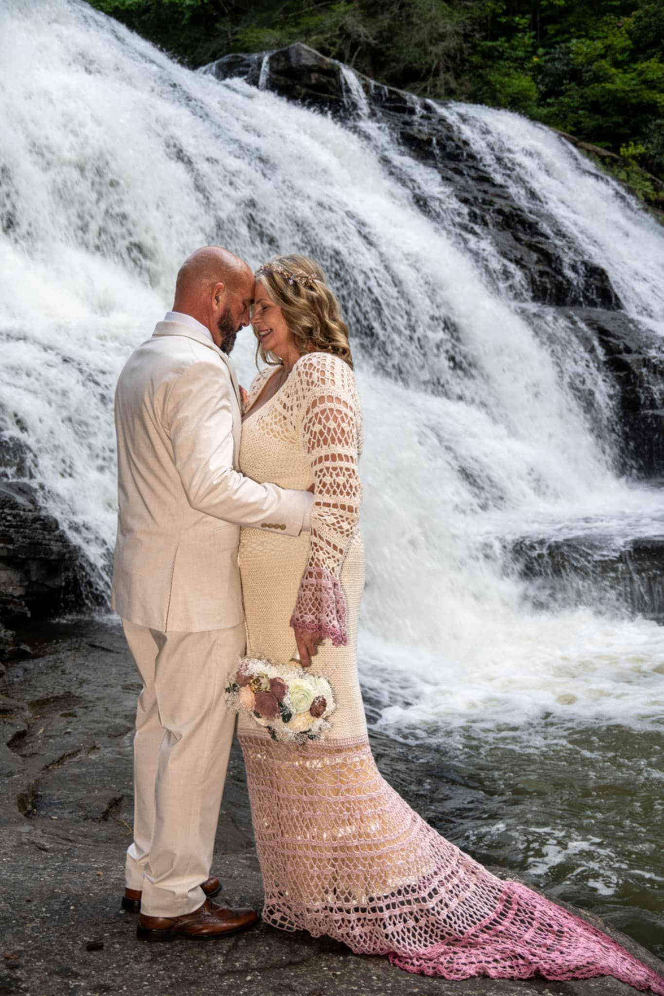 Bride and groom embrace in front of a stunning waterfall, wearing elegant cream wedding attire with a crochet dress.