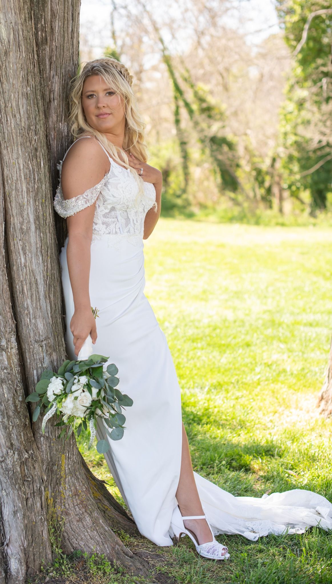 Bride in white off-shoulder lace wedding gown holding bouquet, leaning against tree in lush green outdoor setting.