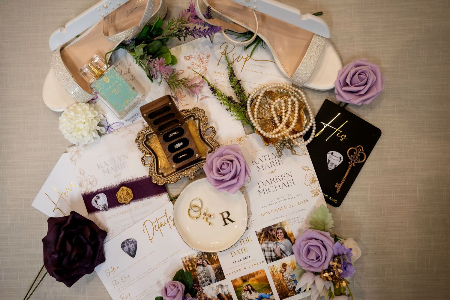 Elegant wedding flat lay with purple roses, pearl jewelry, invitation suite, bridal shoes, and gold accessories on white background.