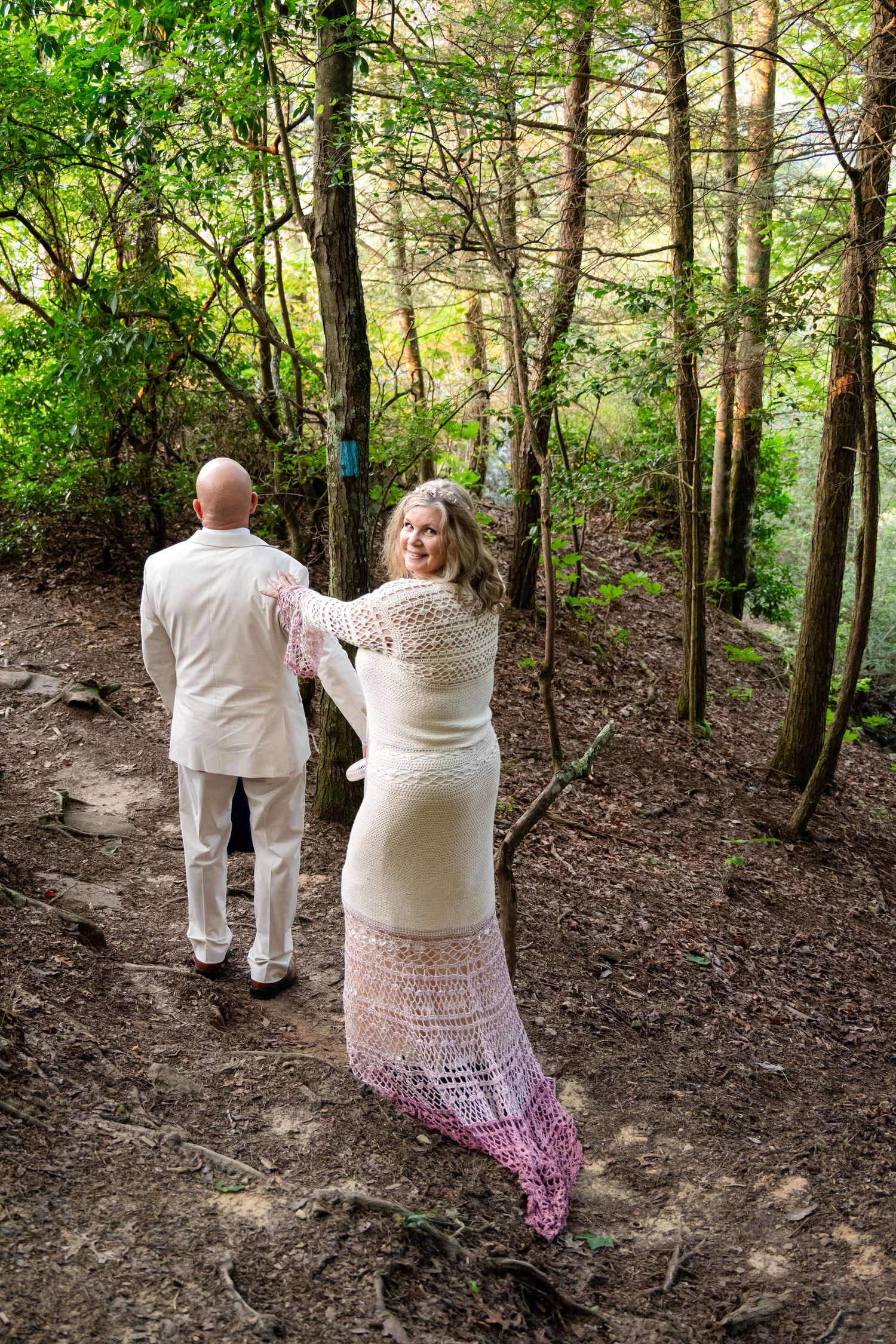 Bride in lace dress and groom in white suit walk hand-in-hand along a wooded forest trail.