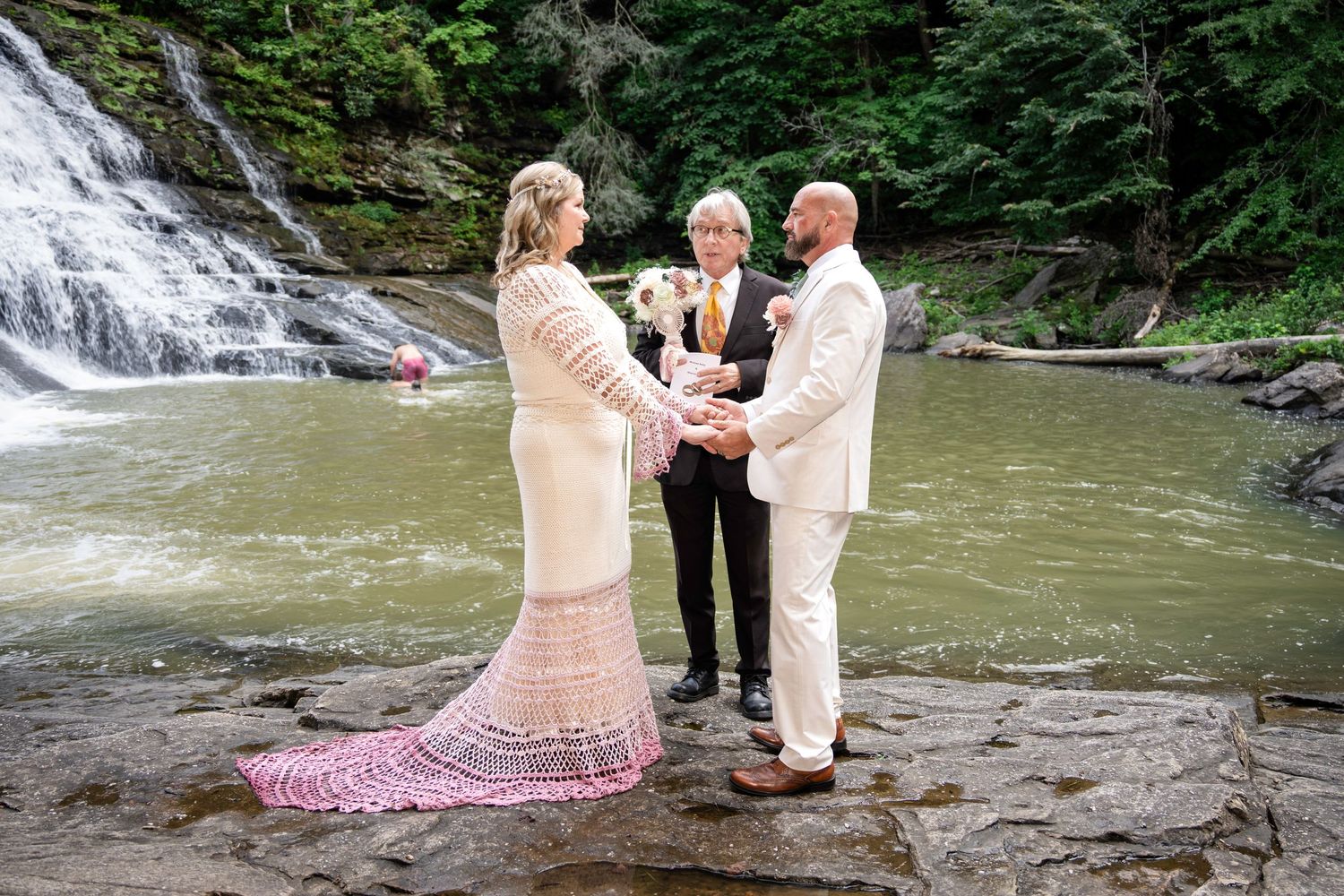 Couple exchanging vows at outdoor waterfall wedding ceremony, bride in lace gown, groom in tan suit, officiant holding flowers.