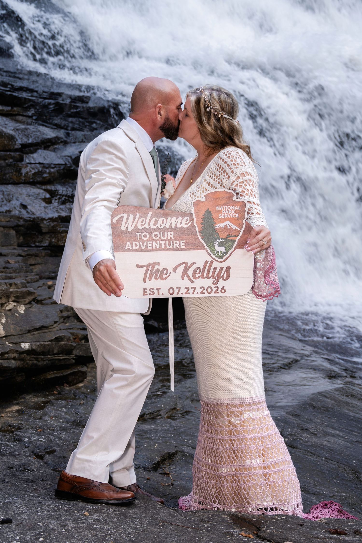 Newlyweds kissing in front of a waterfall holding a welcome sign reading The Kellys Est. 07.27.2026.