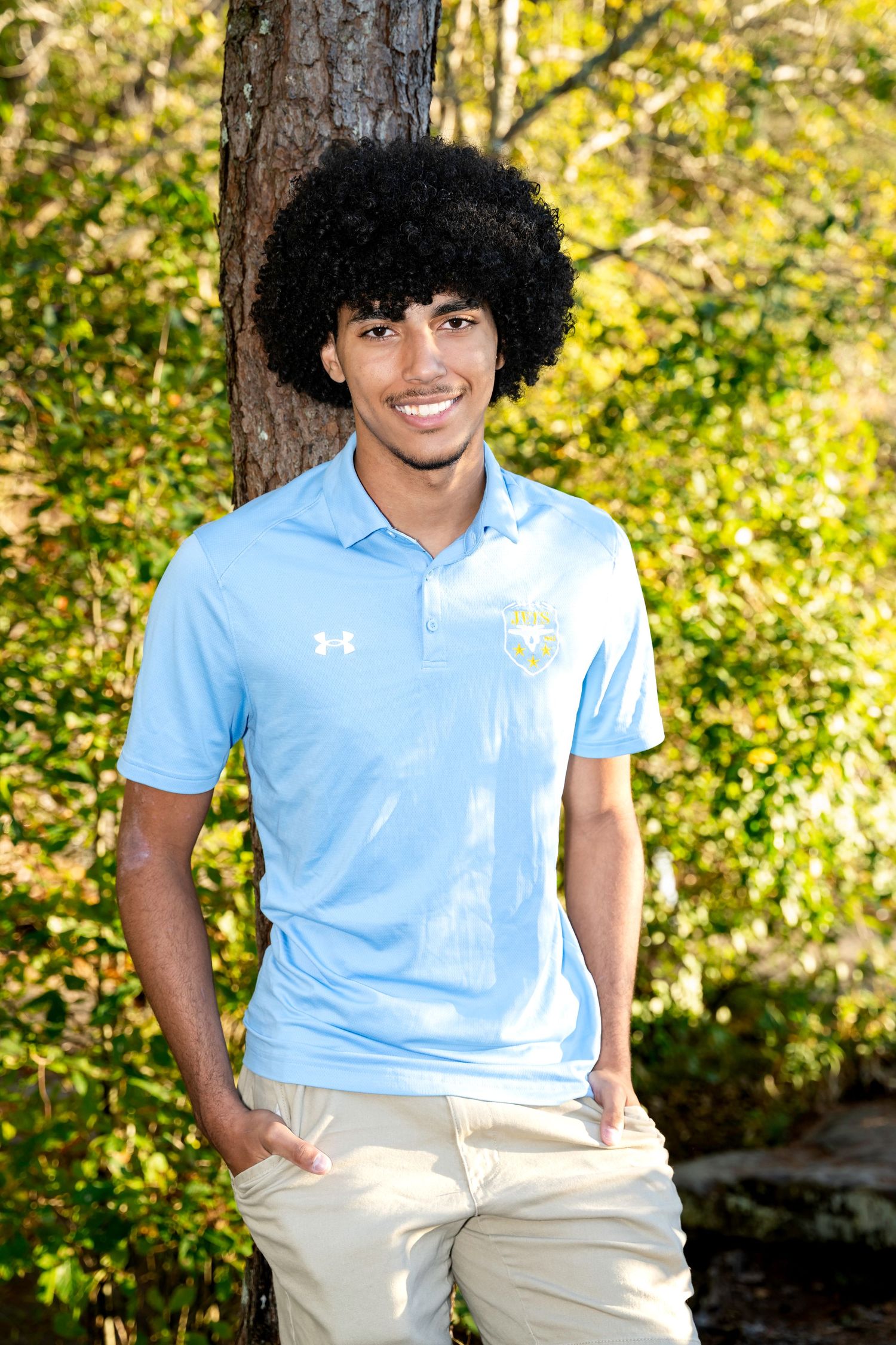 Smiling young man with curly hair wearing a light blue Under Armour polo shirt, standing outdoors near a tree.