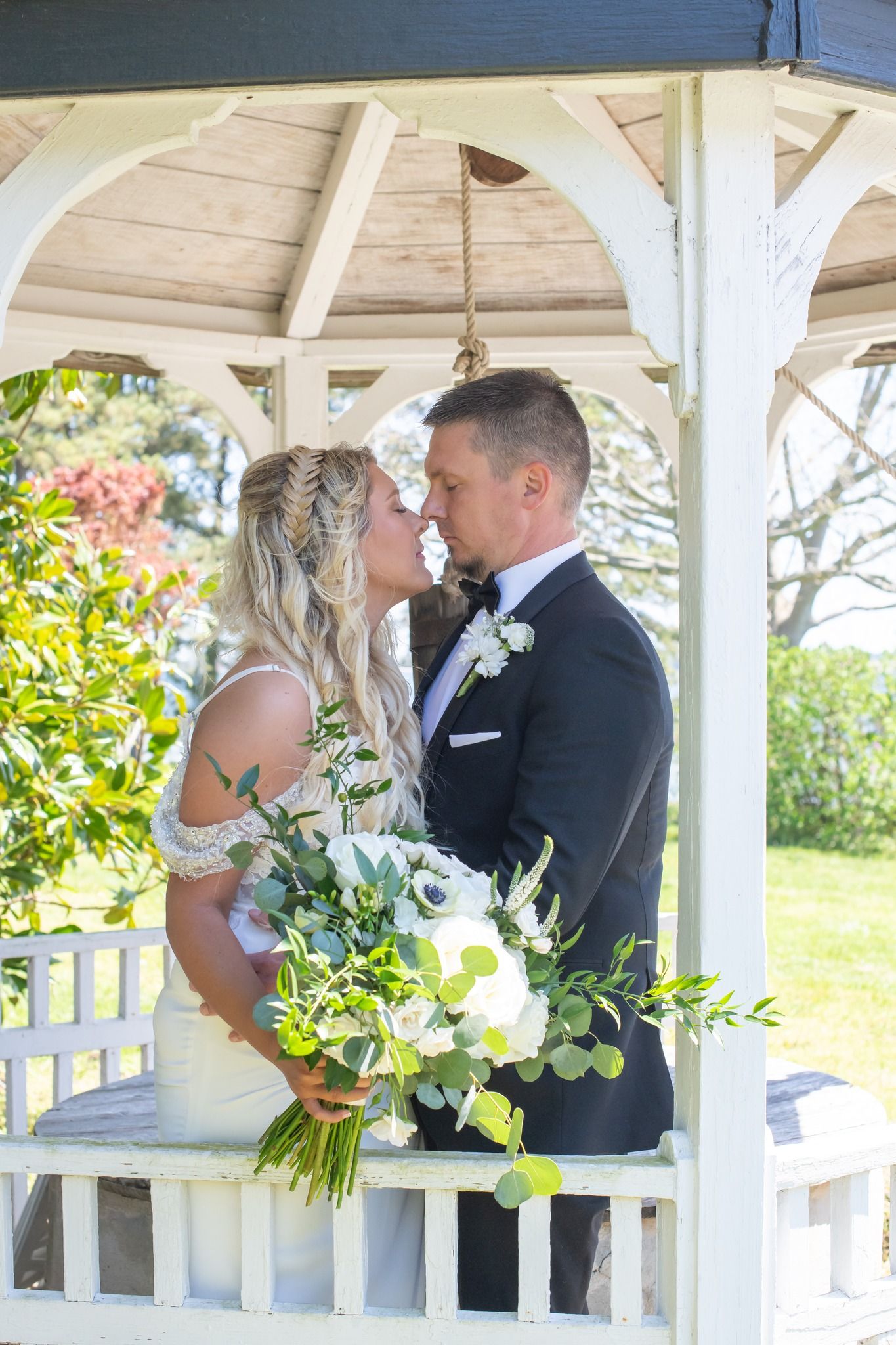 Bride and groom share a tender kiss under a white gazebo with a lush floral bouquet.