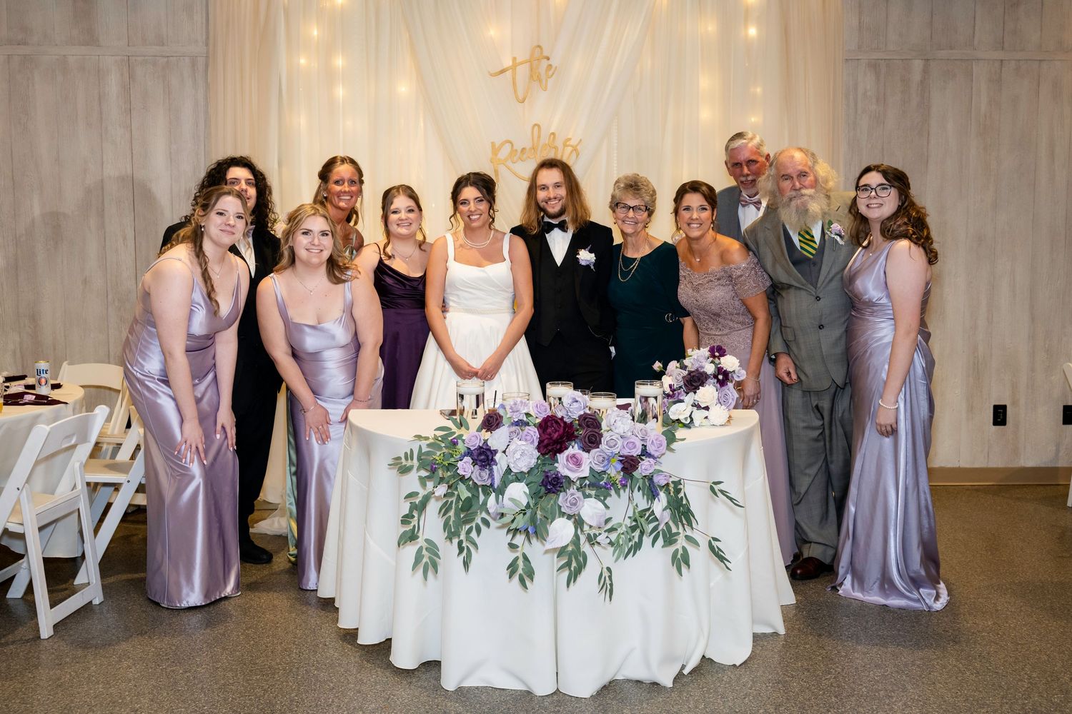 Wedding party group photo with bride, groom, and bridesmaids in lavender gowns at reception.