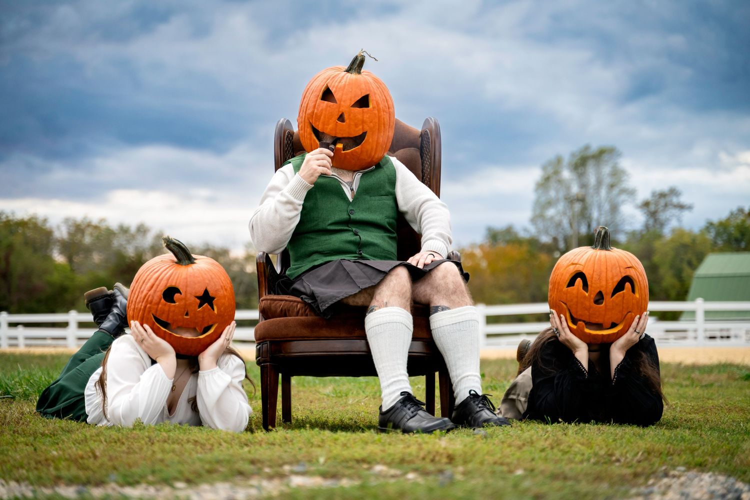 Three people wearing carved jack-o-lantern pumpkin heads pose outdoors in Halloween costumes on a grassy field.