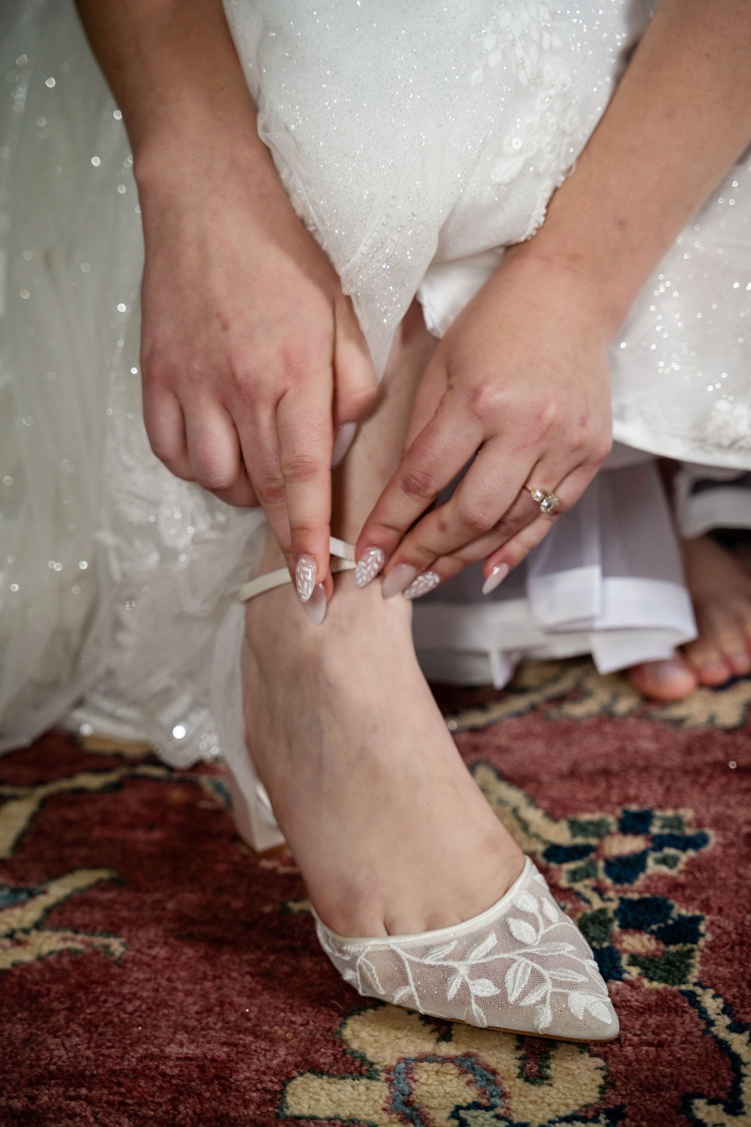 Bride adjusting lace wedding shoe strap with manicured nails on ornate patterned rug.