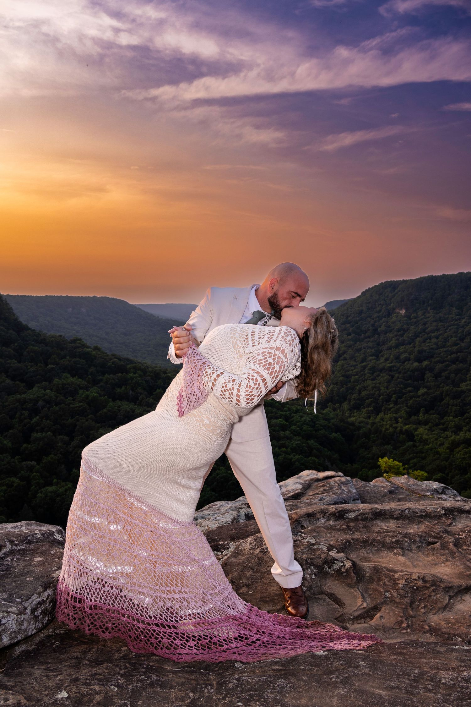 Bride and groom share romantic dip kiss on mountain overlook during stunning golden sunset wedding photo.