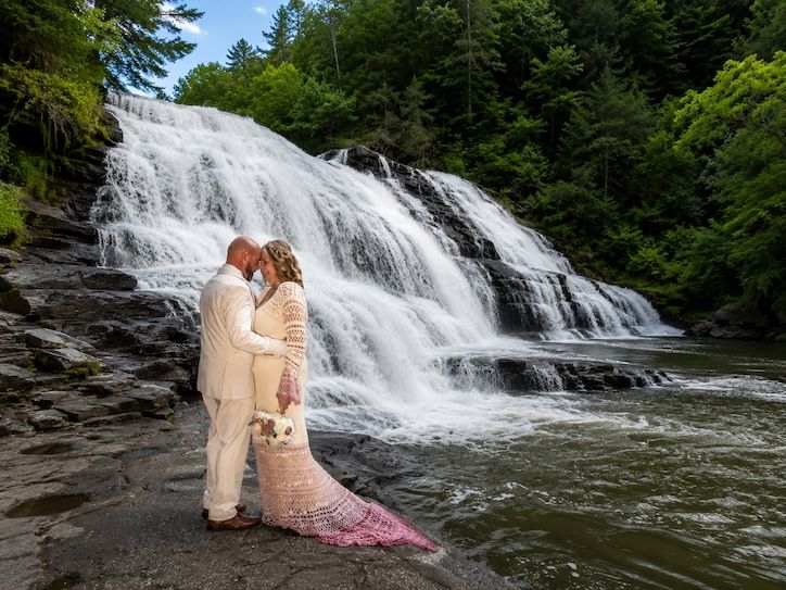 Couple kissing in wedding attire before a stunning cascading waterfall surrounded by lush green forest.