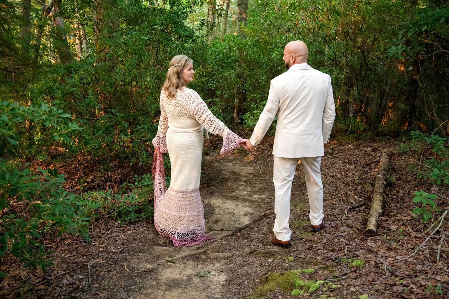 Couple in wedding attire holding hands on a forest path, bride in lace dress with pink shawl, groom in cream suit.
