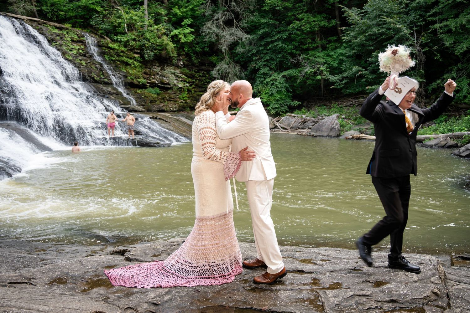 Couple shares a kiss in wedding attire by a waterfall, as a photographer captures the romantic outdoor ceremony.