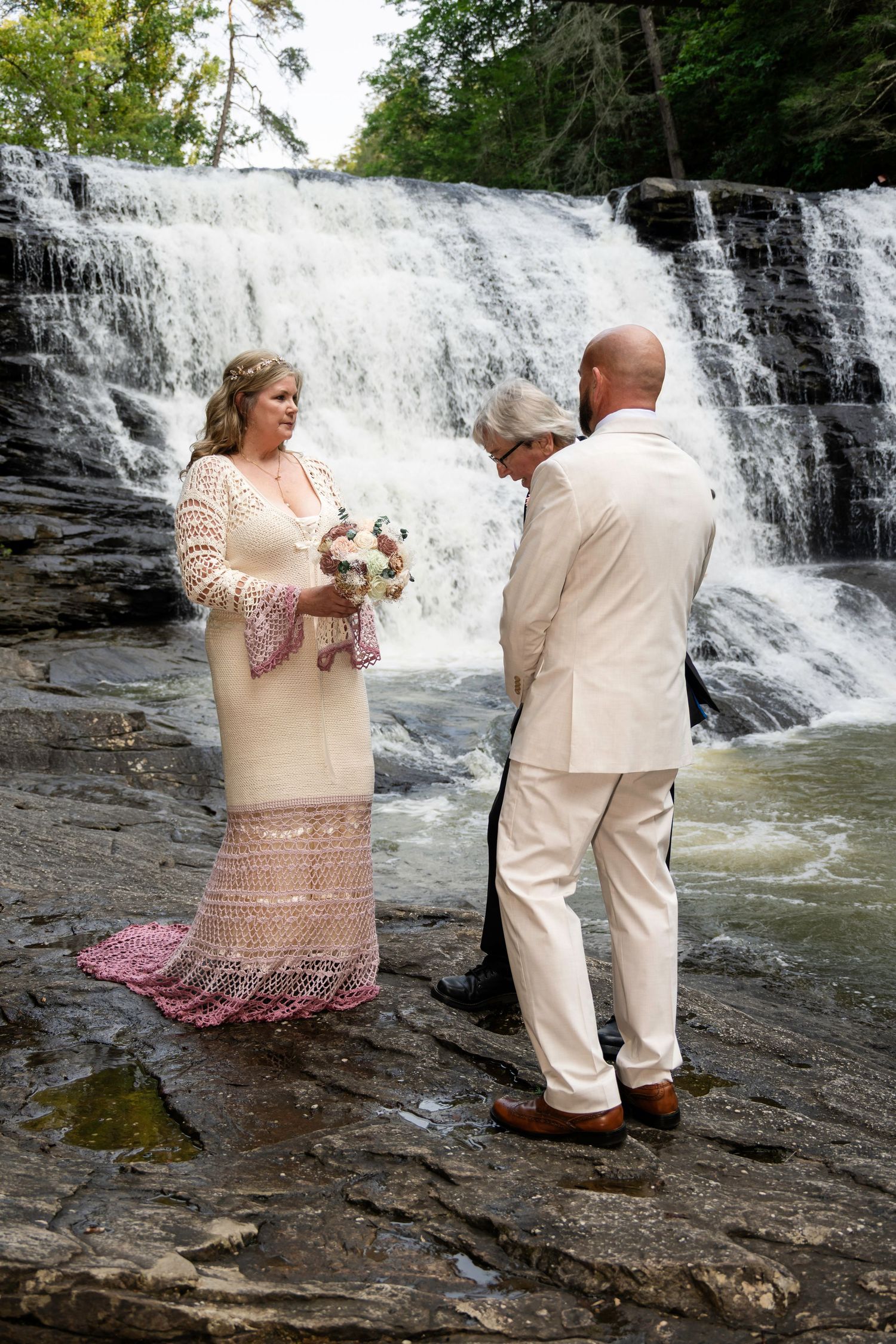Couple exchanges vows before a cascading waterfall, bride in lace dress holding bouquet, officiant presiding outdoors.