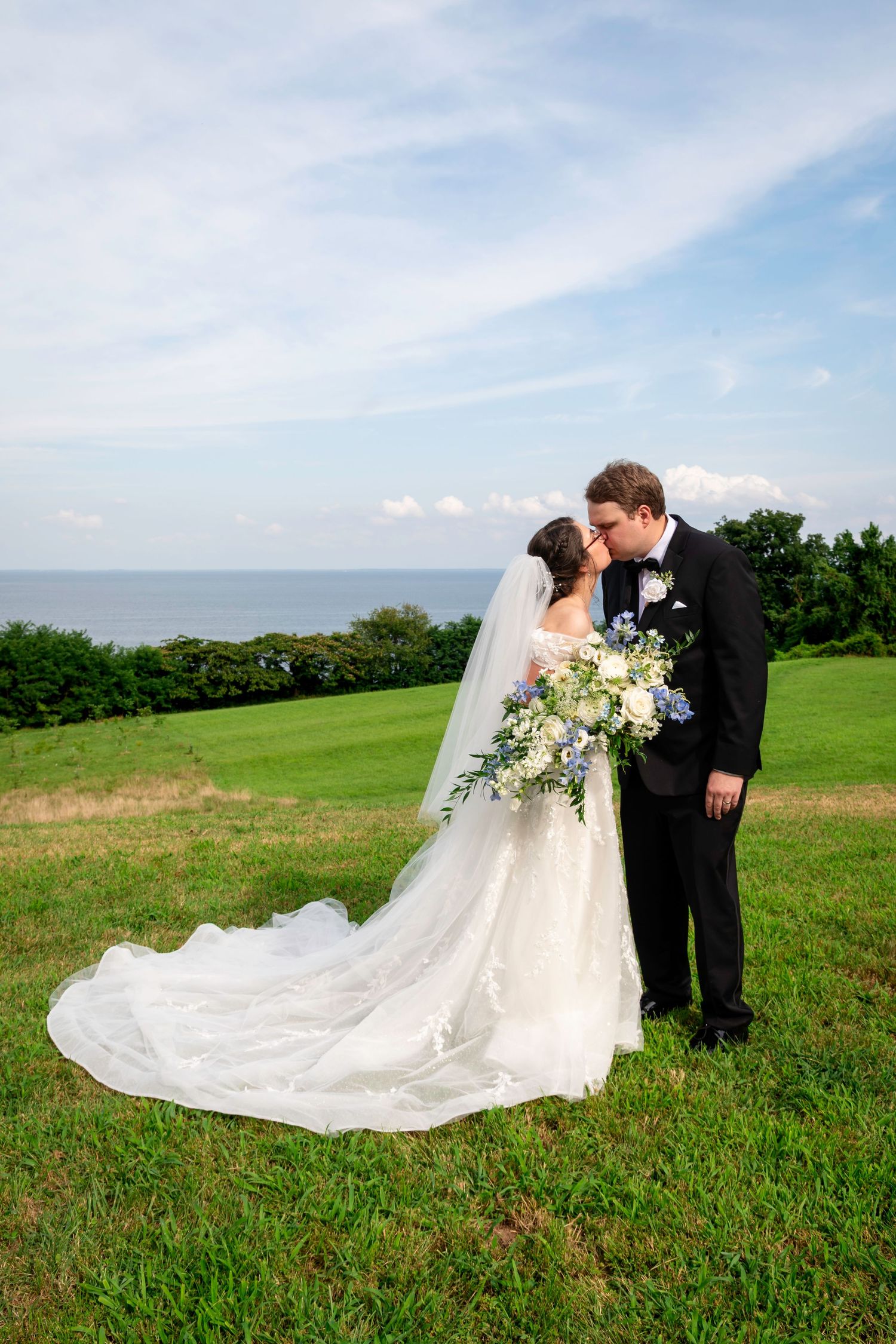 Bride and groom kissing on green lawn with ocean backdrop, bride wearing long veil and holding blue and white bouquet.