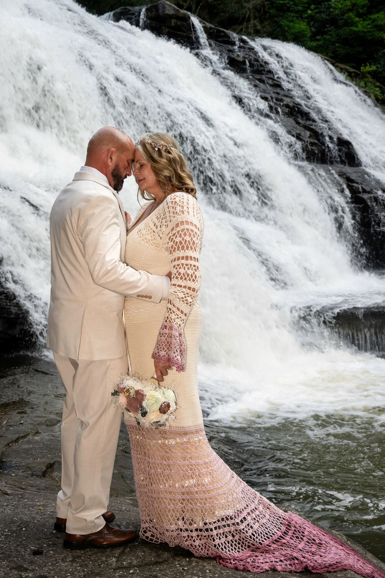 Bride and groom share a romantic moment in front of a stunning waterfall, bride wearing a crochet wedding dress.