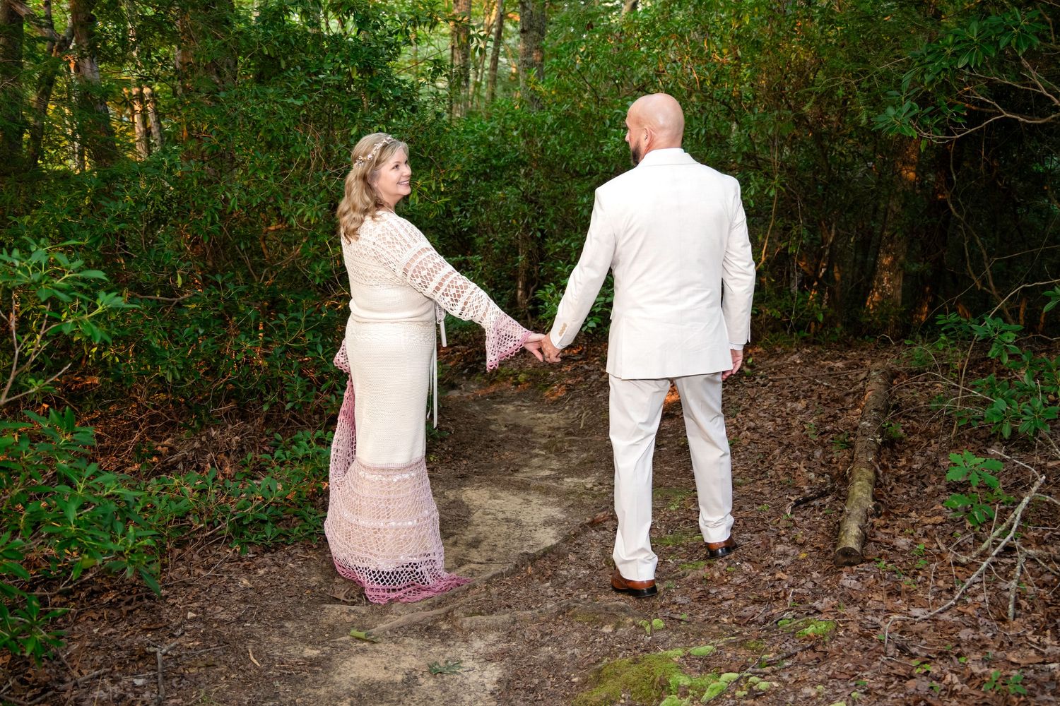 Couple in formal attire holding hands on a forest path, woman in floral gown, man in white suit.