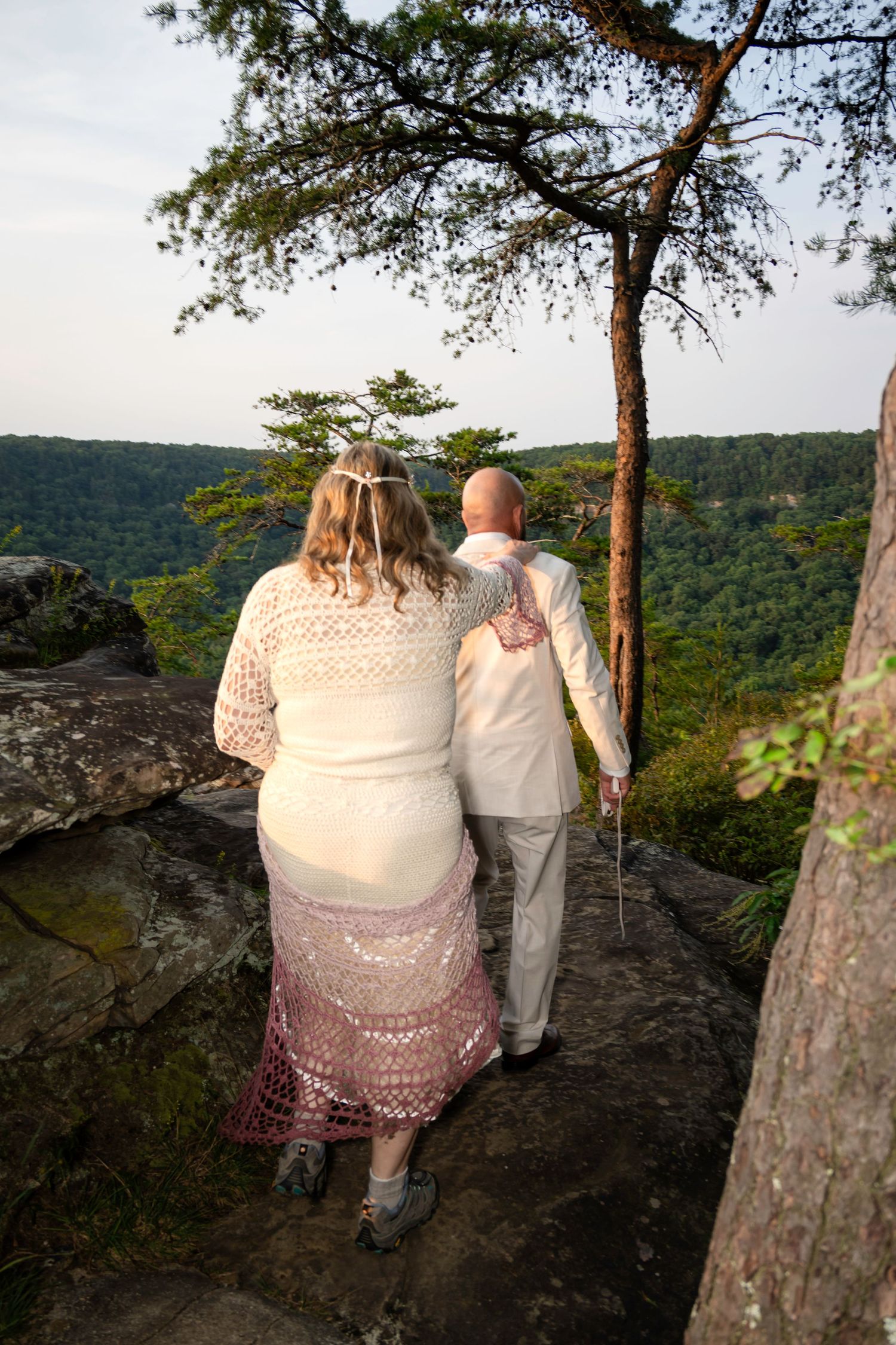 Couple in wedding attire standing on rocky overlook surrounded by lush green forest landscape at sunset.
