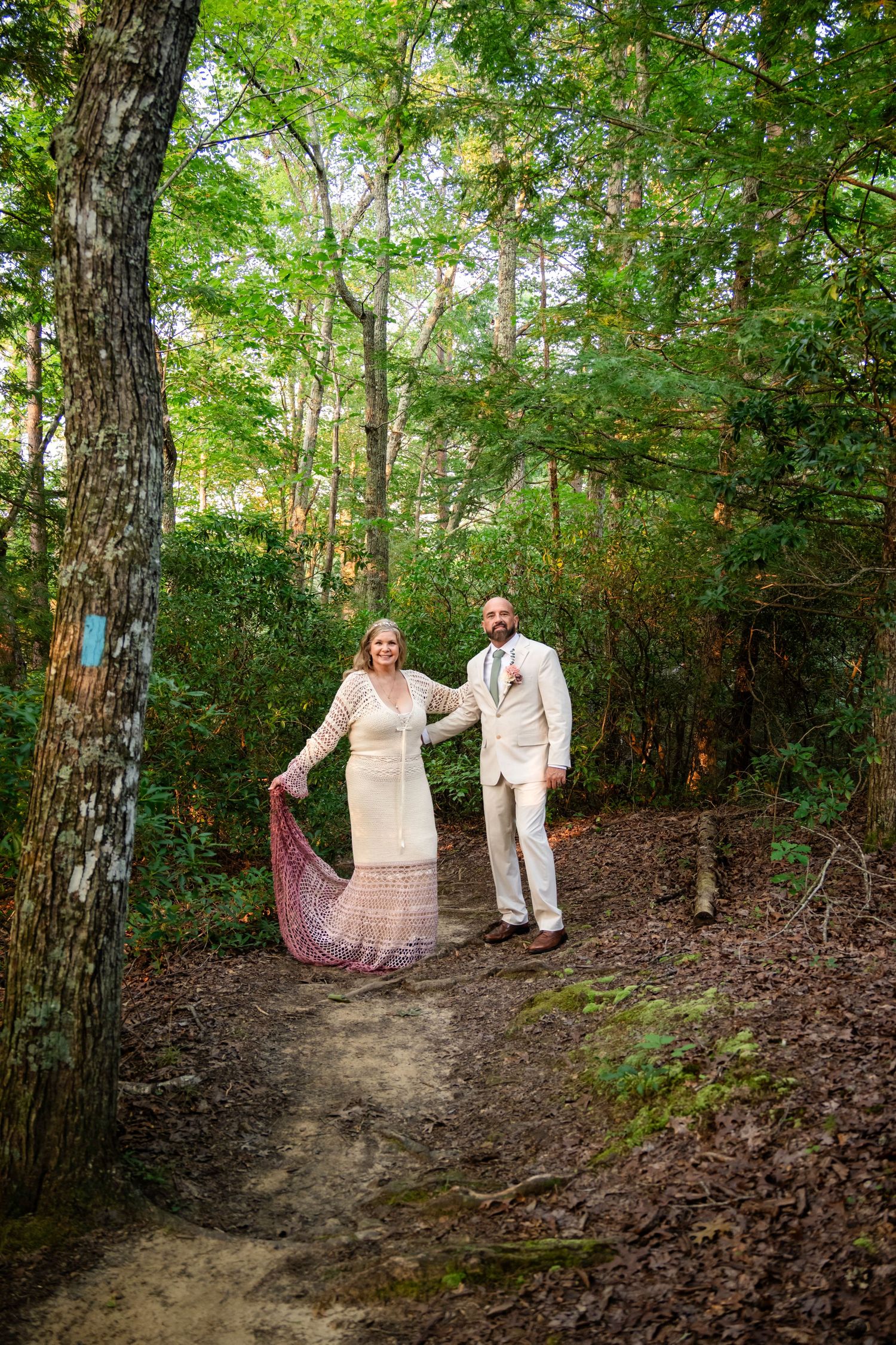 A bride and groom hold hands on a forest trail, she in a lace dress with red shawl, he in a cream suit.