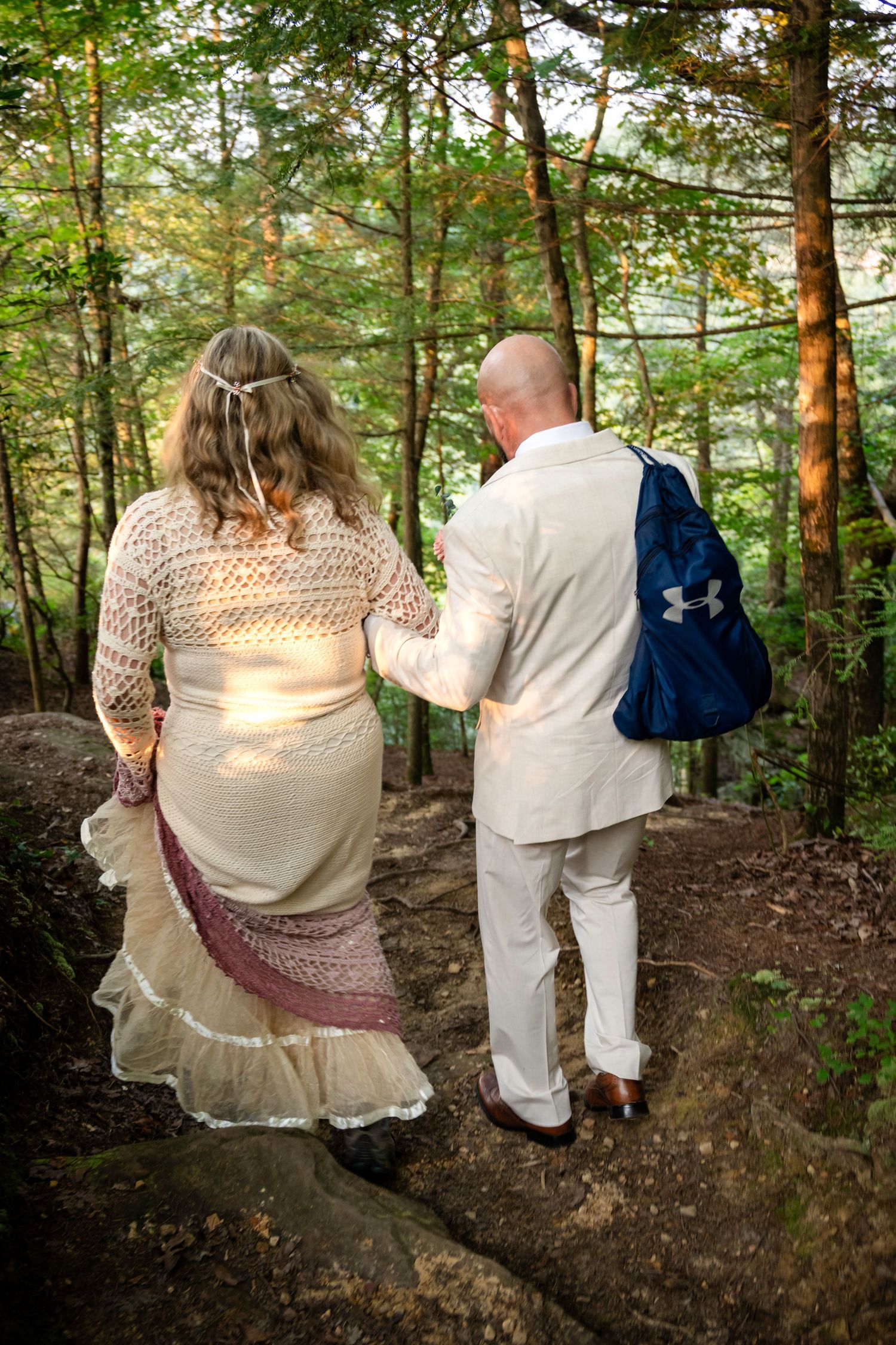 A couple in formal attire walks hand-in-hand through a lush forest, the woman in a boho dress and the man in a white suit.
