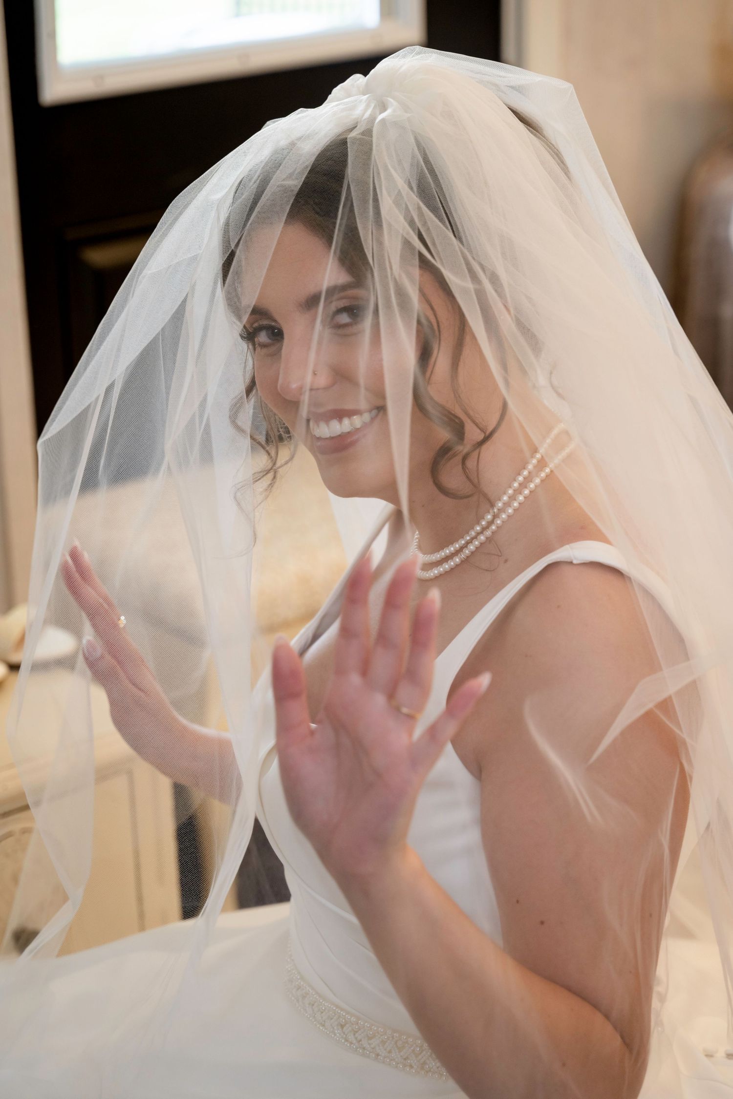 Smiling bride in white veil and pearl necklace playfully posing with hands raised.