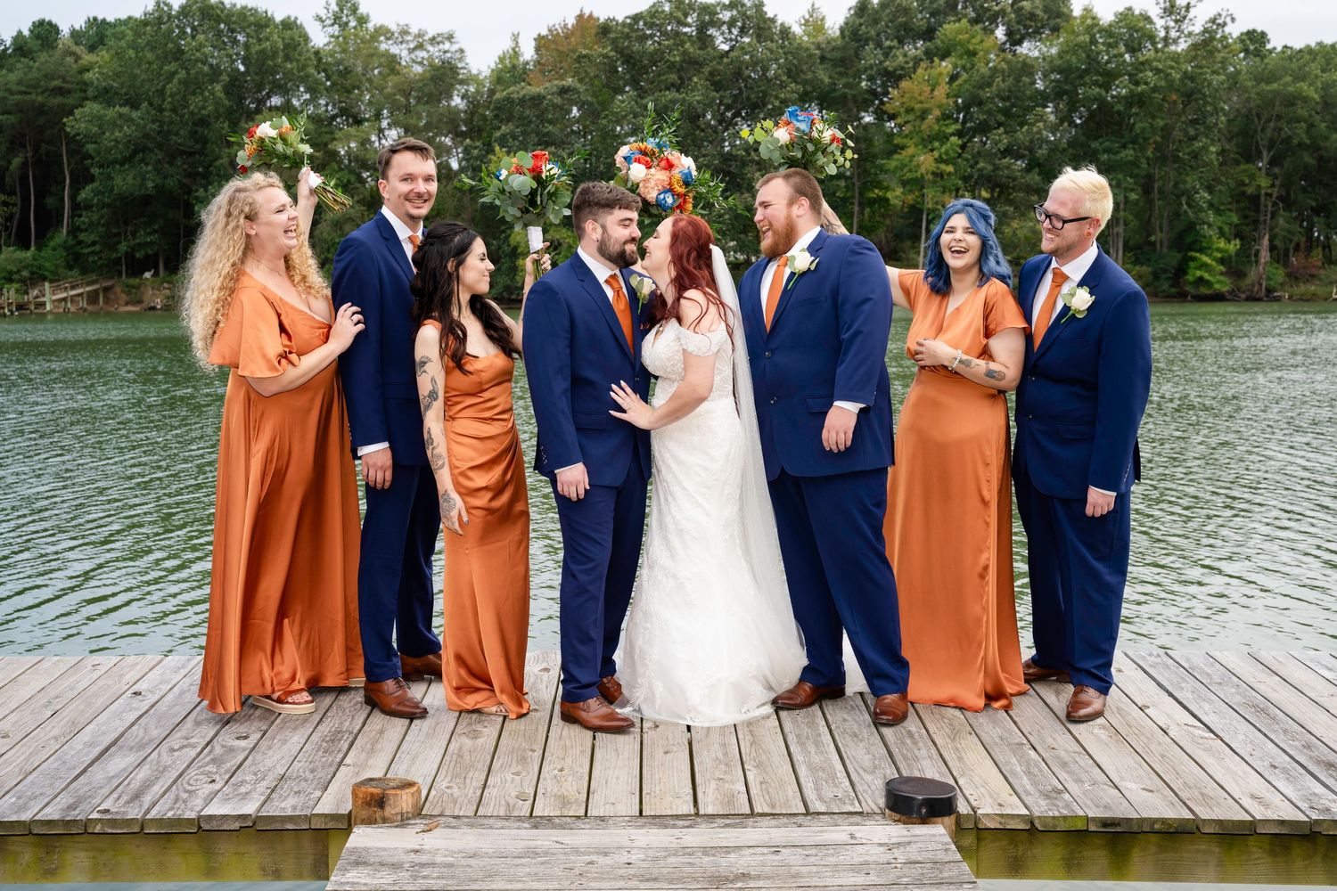 Wedding party poses on dock by lake, bridesmaids in burnt orange gowns and groomsmen in navy suits flanking bride and groom.