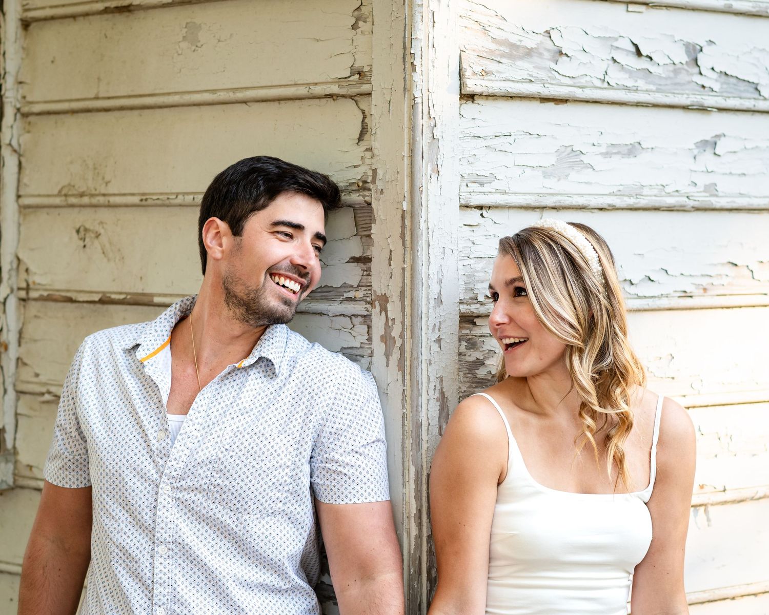 A smiling couple leaning against a rustic white wooden wall, enjoying a casual outdoor moment together.