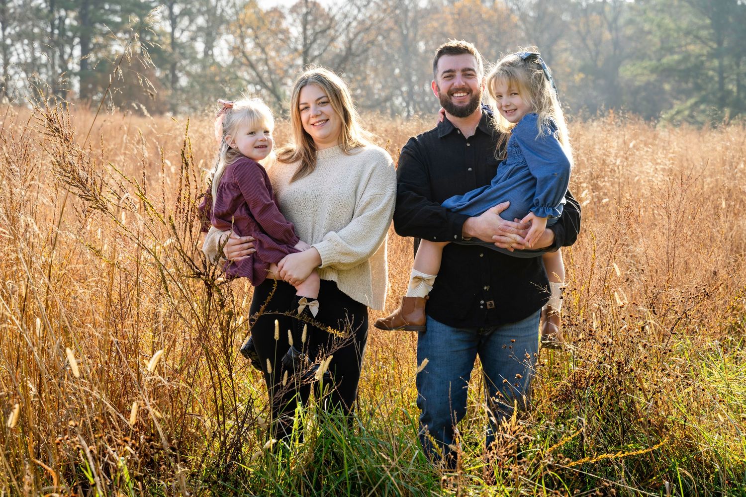 Family of four posing in golden autumn field, parents holding two young children, smiling outdoors in warm fall sunlight.