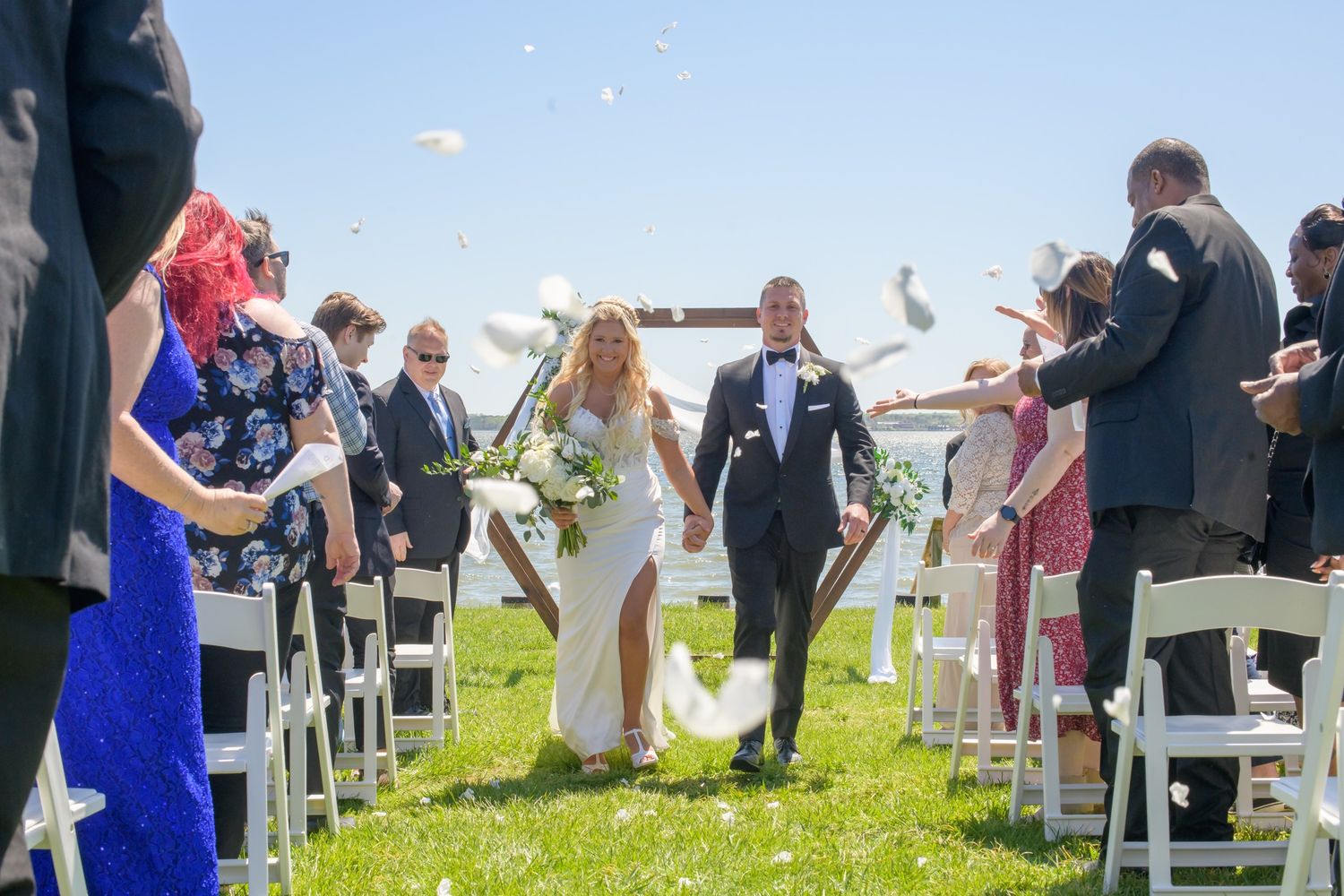 Newlyweds walking down aisle as guests toss white petals at sunny outdoor waterfront wedding ceremony.
