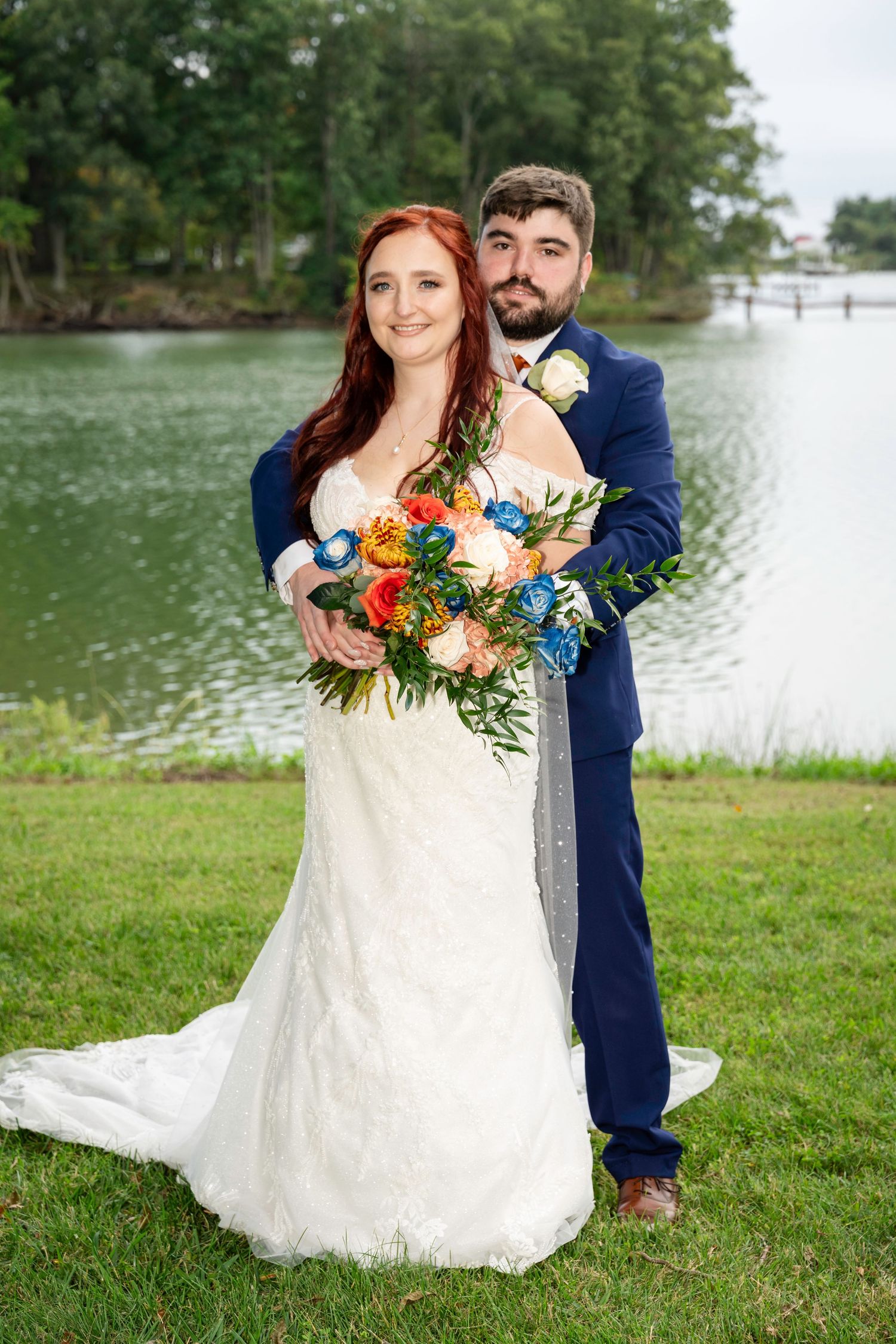 Bride and groom pose together by a serene lake, she holding a vibrant bouquet of blue, orange, and peach flowers.