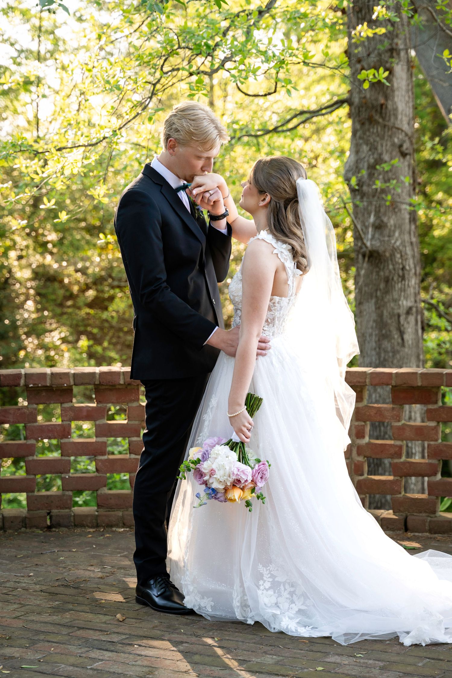 Groom in black tux kisses bride's hand outdoors, bride holds colorful bouquet in white lace gown with veil.