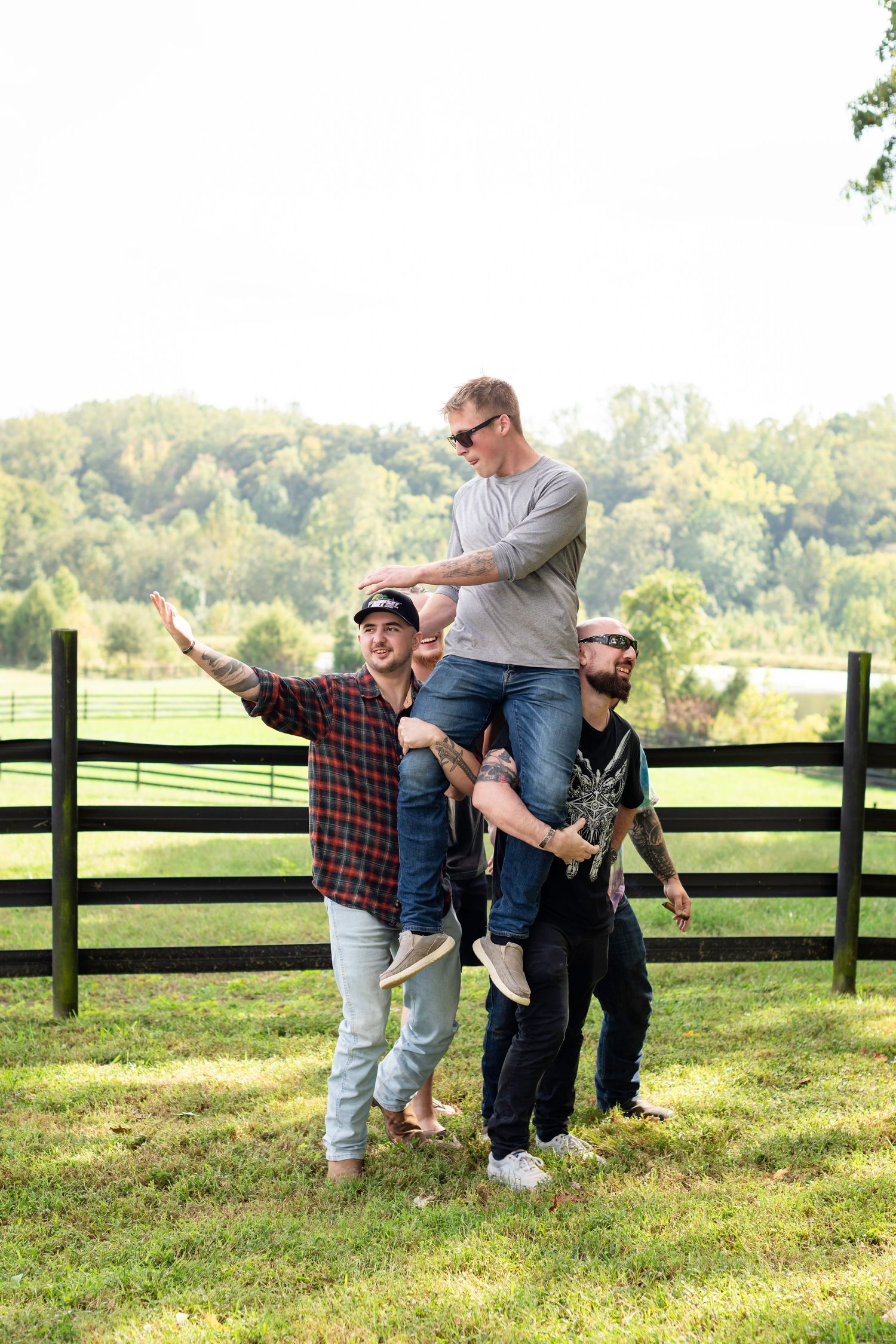 Four men posing outdoors near a wooden fence, one sitting on shoulders, on a sunny day in a green countryside setting.