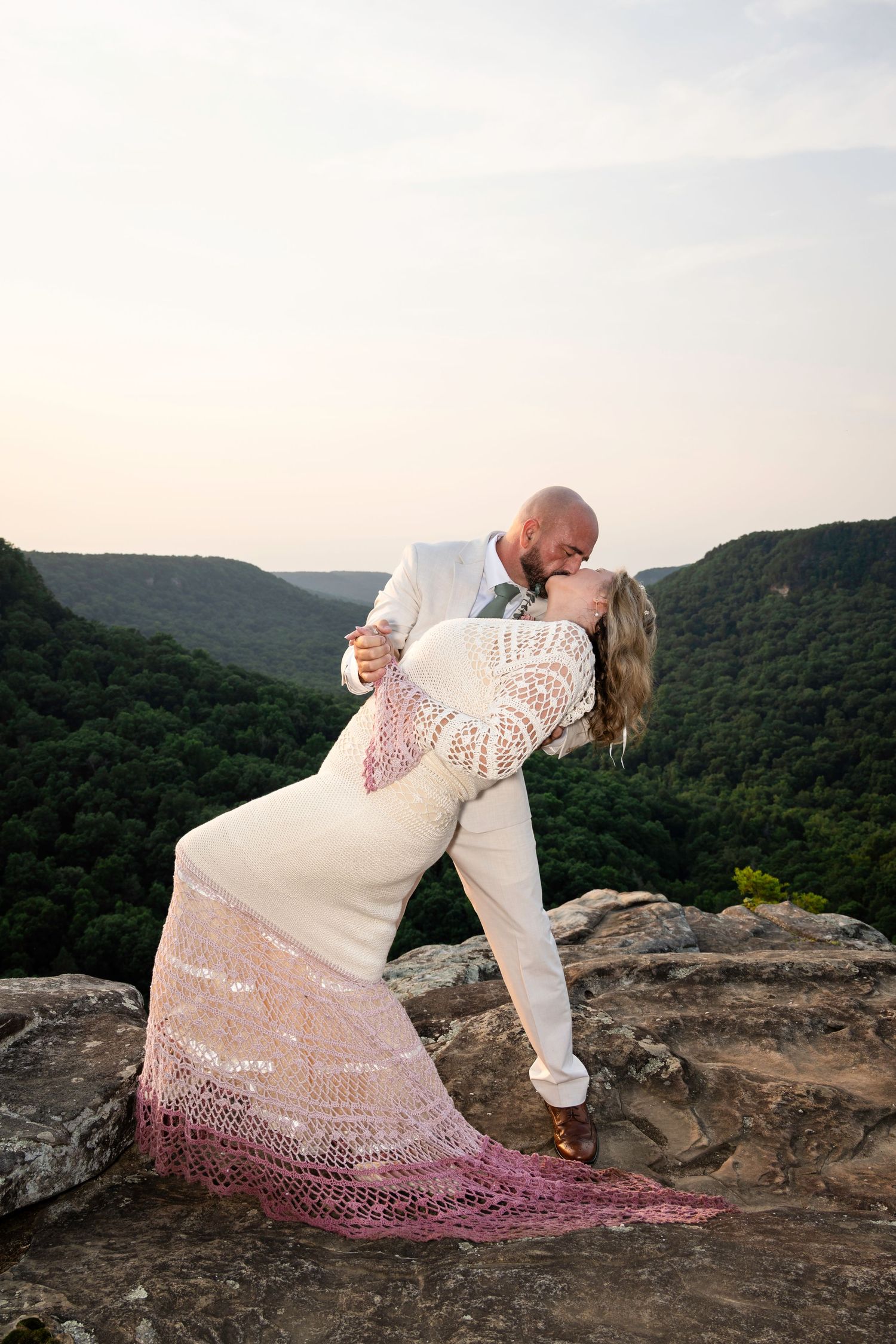 Couple sharing a romantic dip kiss on a rocky overlook with lush green mountain valleys at sunset.