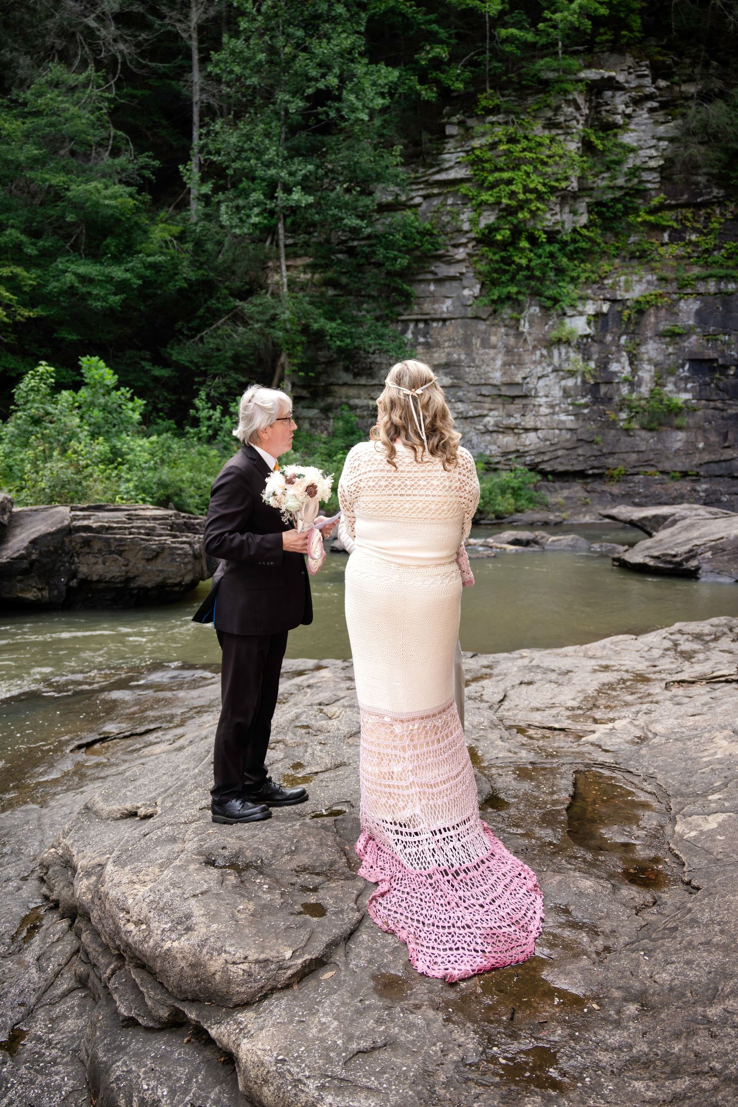 Bride in pink crochet gown holds bouquet with groom in black suit on rocky riverbank surrounded by lush forest.