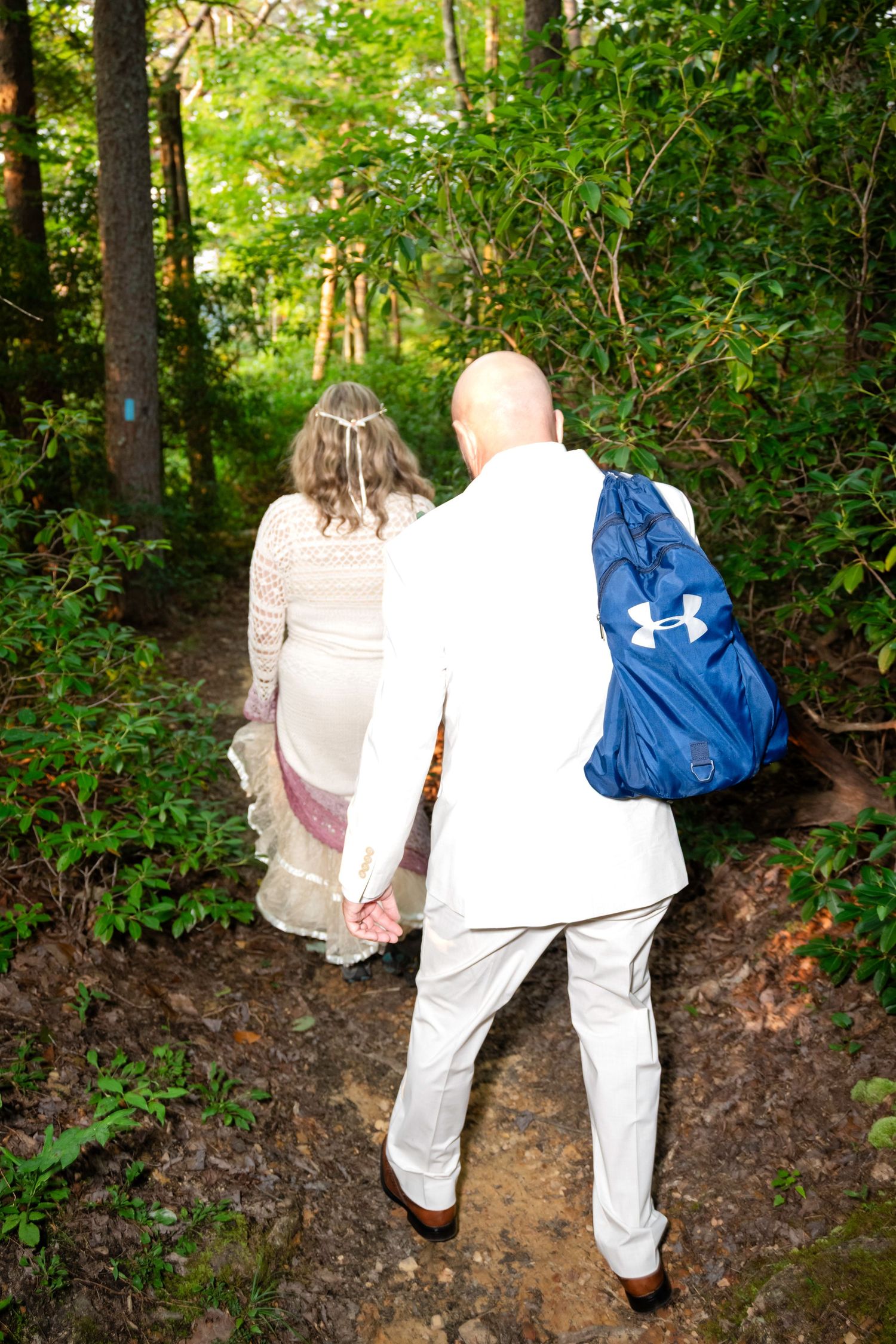 Bride and groom walking hand in hand down a forest trail, groom wearing white suit with blue Under Armour backpack.