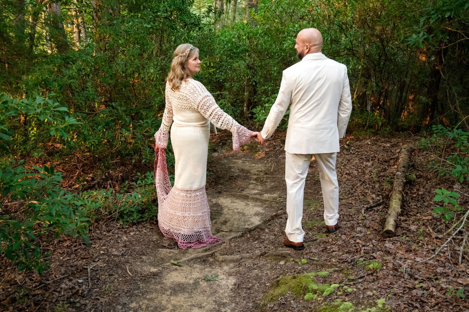 Couple in wedding attire holding hands on a forest path surrounded by lush green trees.