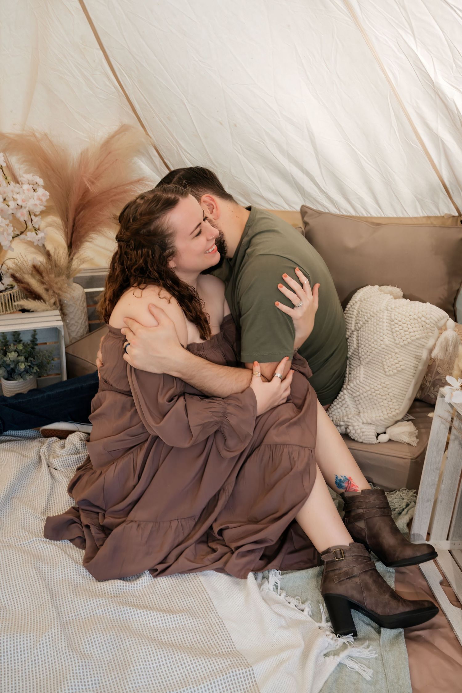 Couple sharing a romantic embrace inside a boho-styled tent with neutral decor, pillows, and pampas grass.