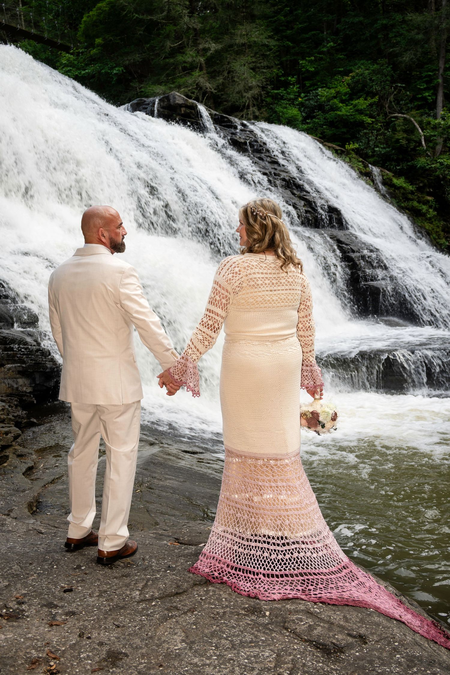 Bride and groom hold hands facing a cascading waterfall, she wears a lace ombre gown holding a bouquet.