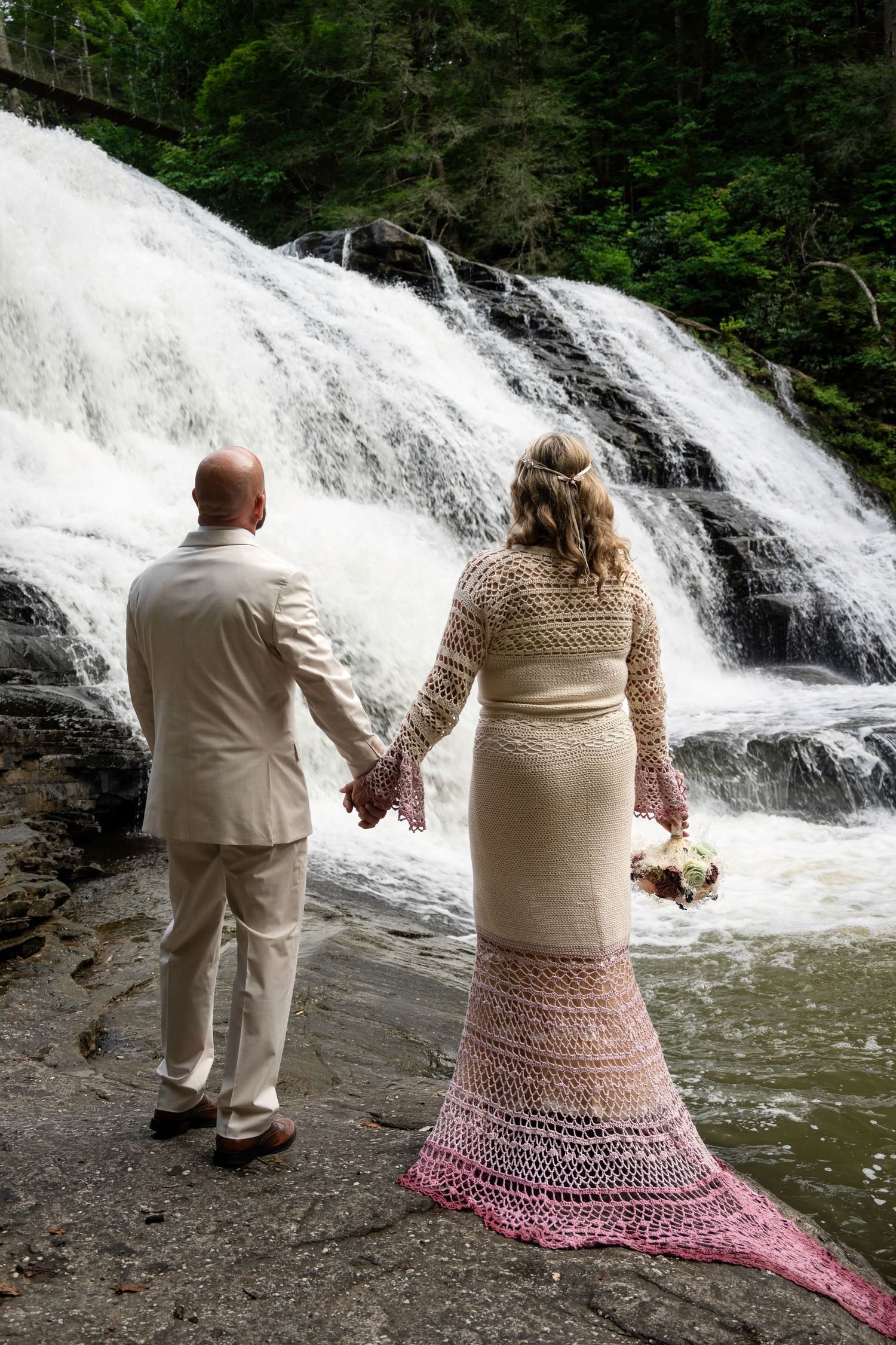 Bride and groom hold hands facing a cascading waterfall, she wears a crochet ombre wedding gown with pink hem.